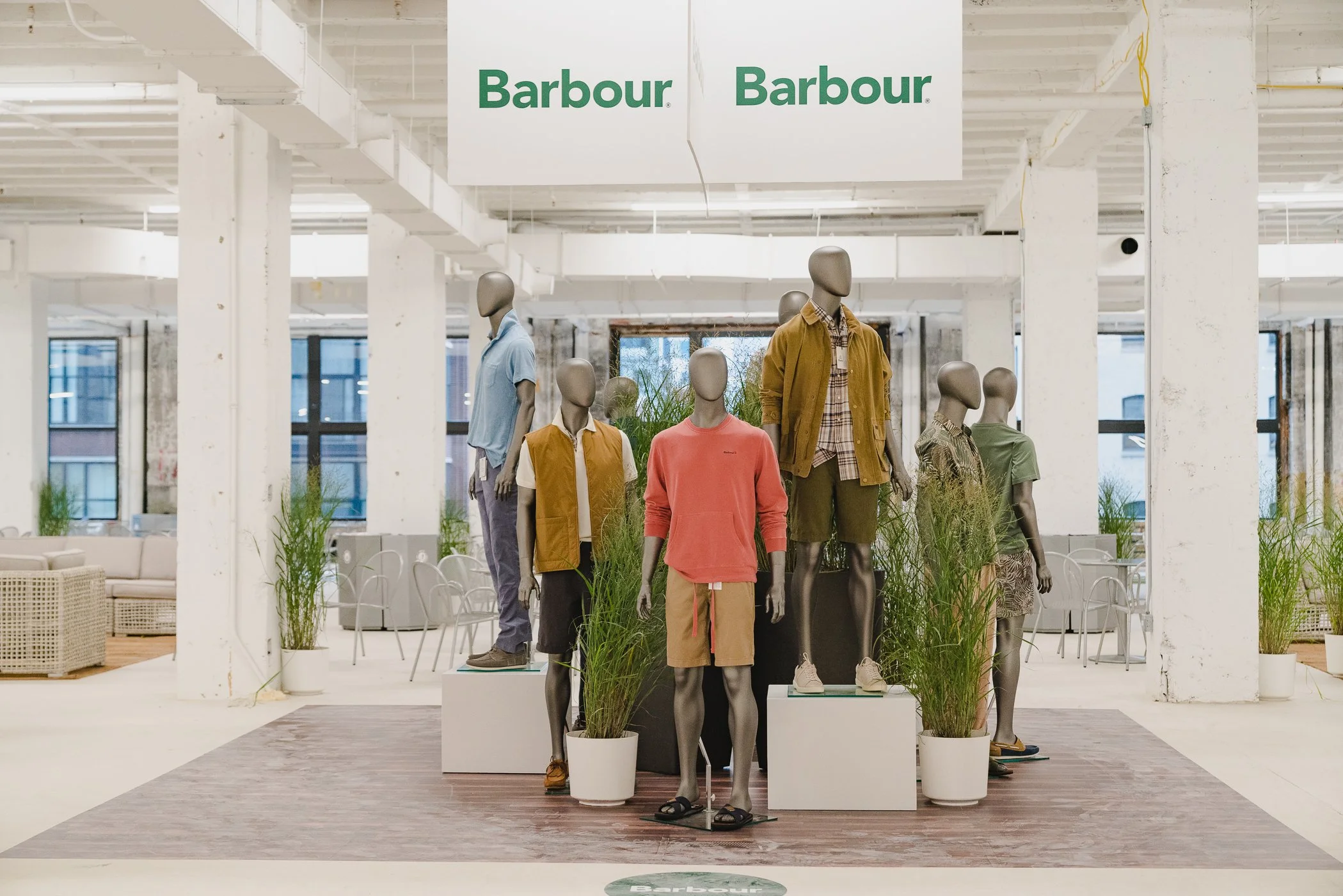 Mannequins dressed in casual clothing inside a store with white pillars and plants, with a large 'Barbour' sign hanging from the ceiling.