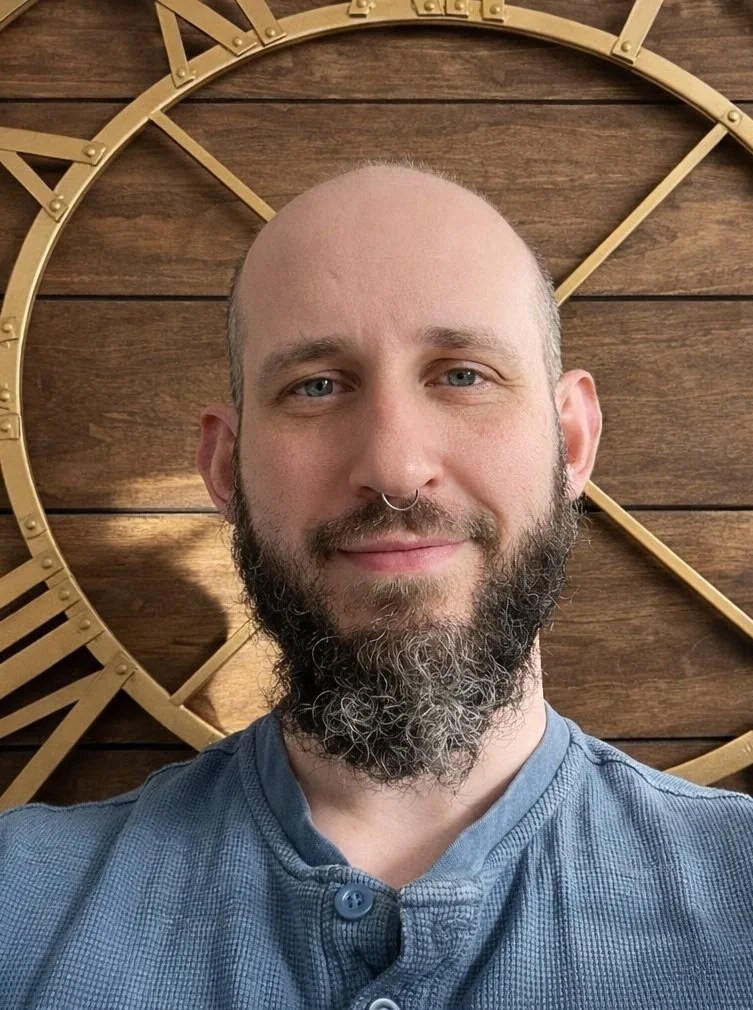 A man with a beard and septum piercing smiling in front of a large clock on a wooden wall.