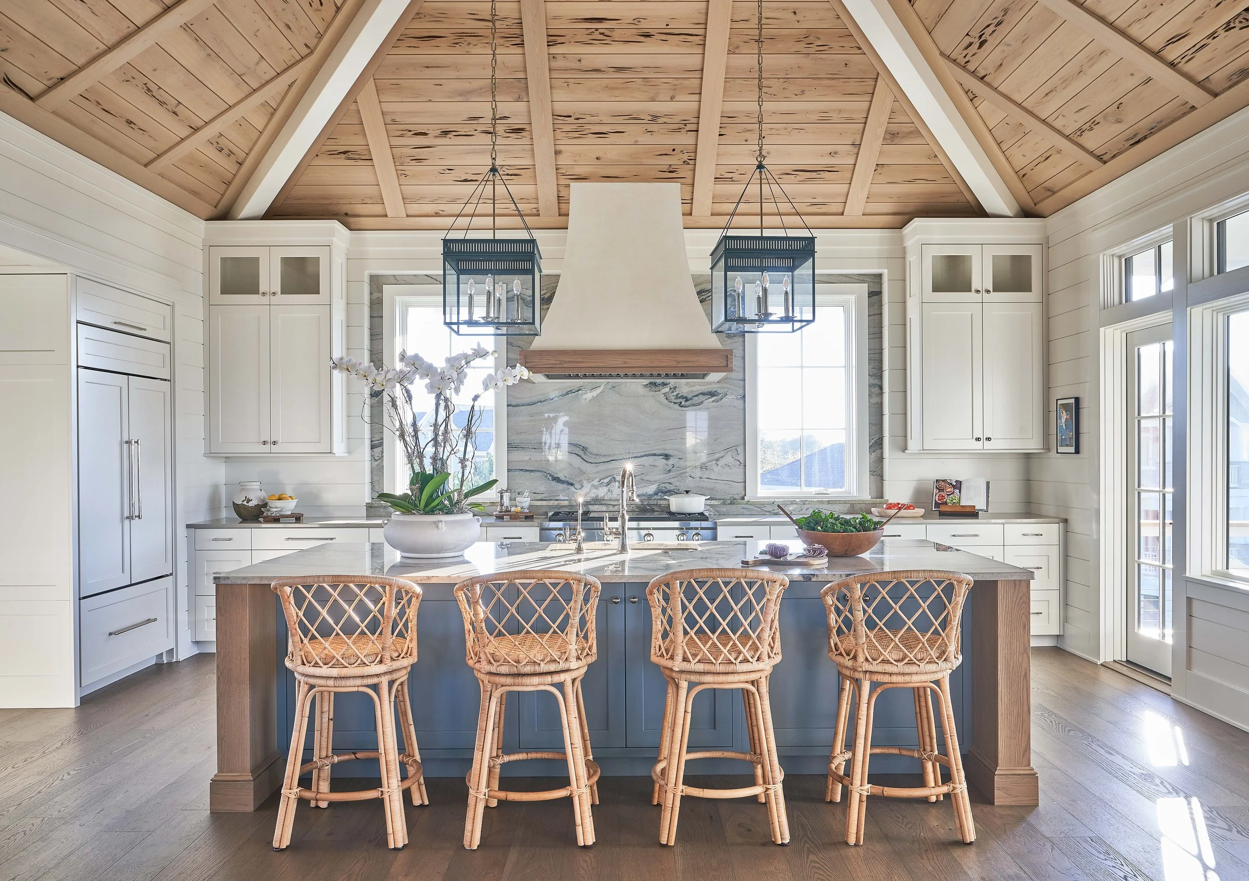 Modern kitchen with white cabinets, a wooden island with four rattan chairs, a marble backsplash, and a wooden vaulted ceiling with two black lantern pendant lights.