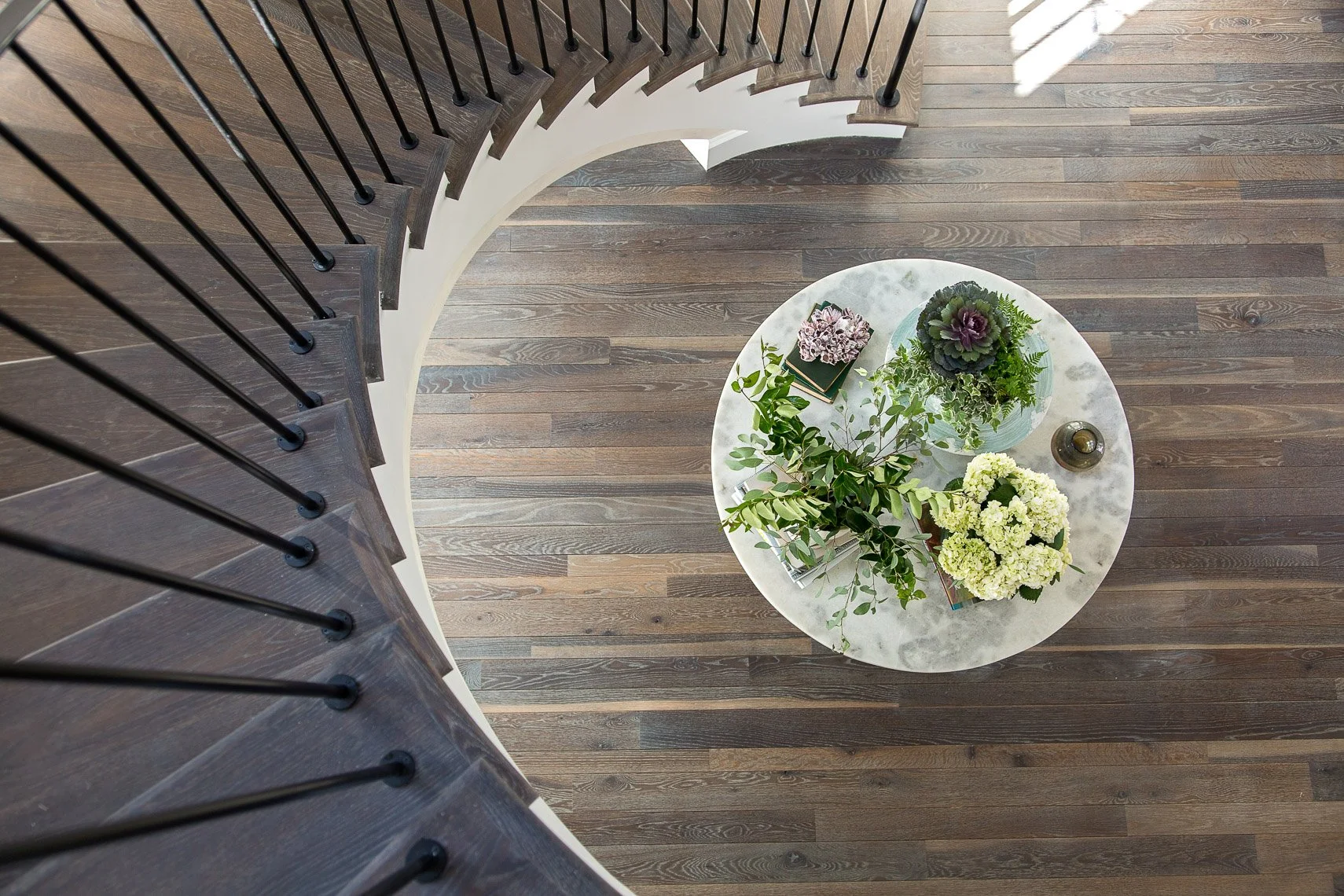 View looking down at a round white marble table with various potted plants, a small decorative object, and some books, from the top of a curved wooden staircase with black balusters.