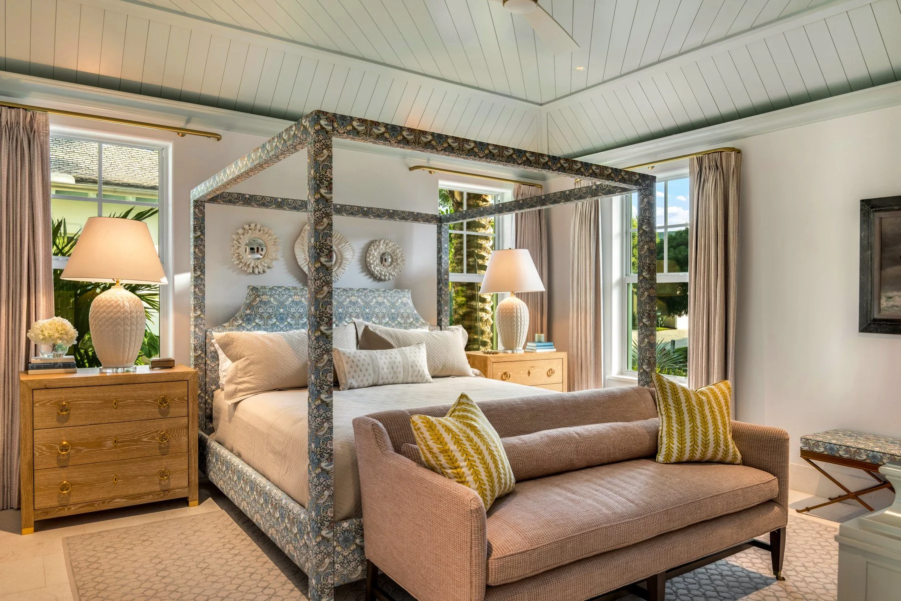 A cozy bedroom with a four-poster bed and patterned bedding, flanked by wooden nightstands with large white lamps, and a pink sofa with yellow and white patterned pillows in front of the bed, with large windows and draped curtains in the background.