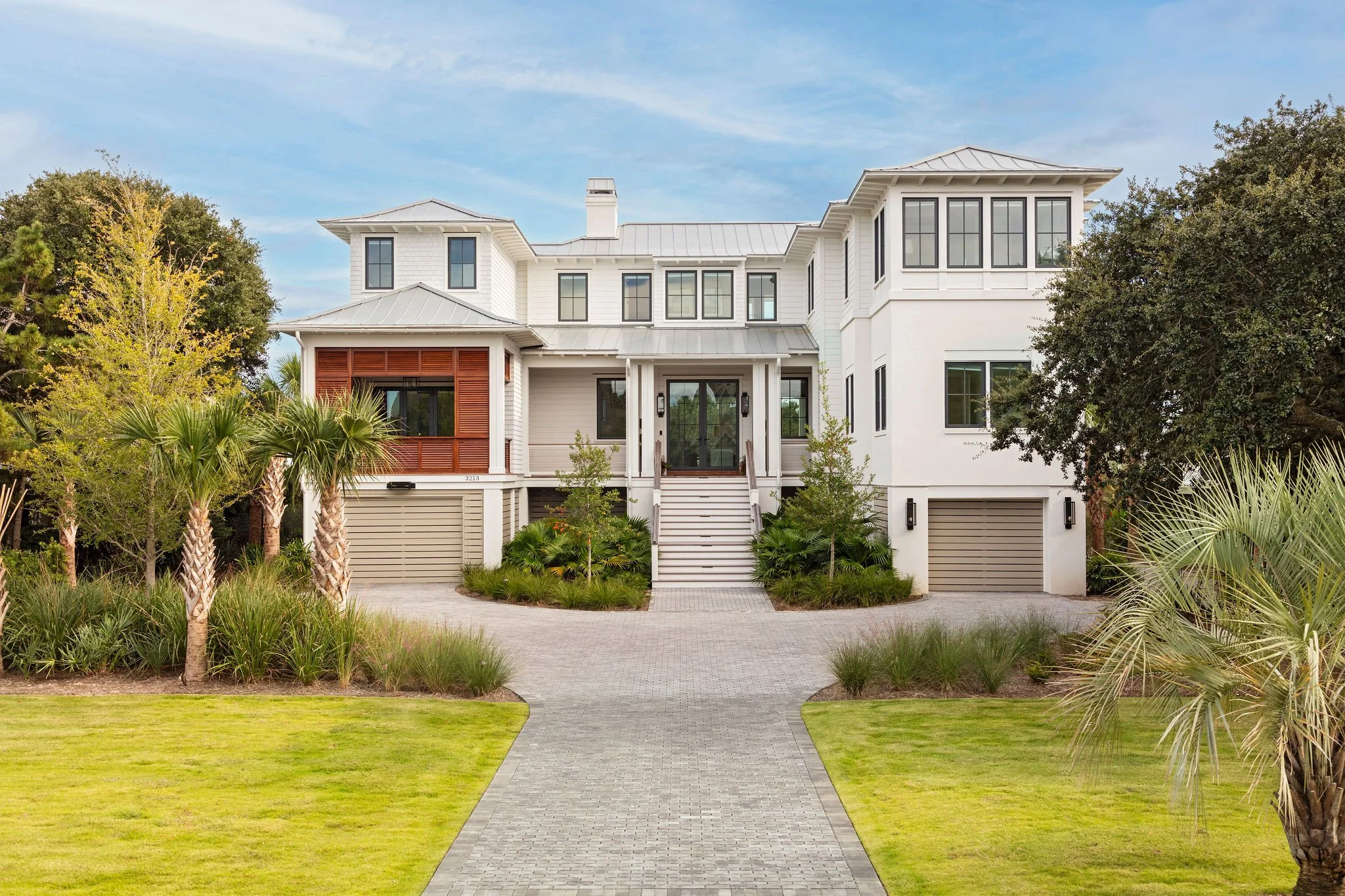 Large modern white house with a gray metal roof and garage doors, surrounded by well-maintained green lawn and landscaping, with palm trees and shrubs, under a blue sky.