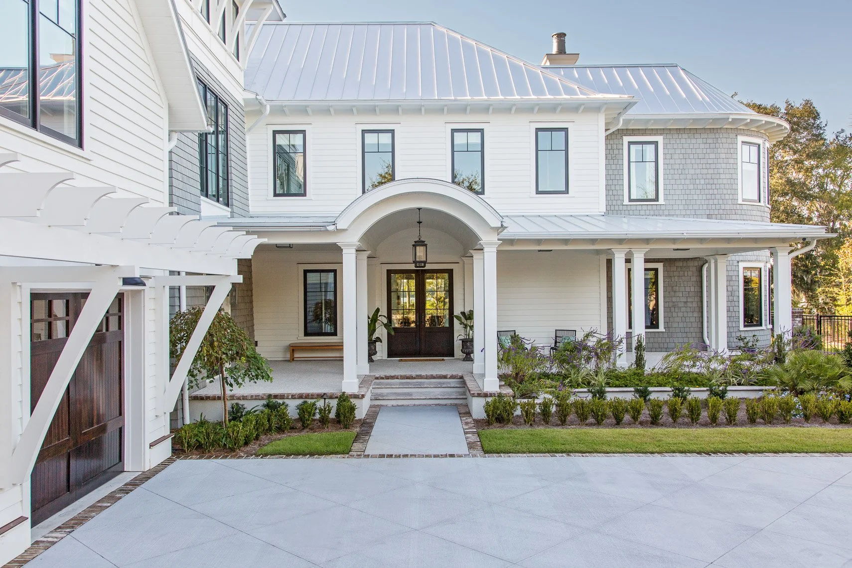 Front view of a large, modern white house with a covered porch and a garden in front. The house has multiple windows, a metal roof, and a mix of siding and shingles. There are trees and a fence in the background.
