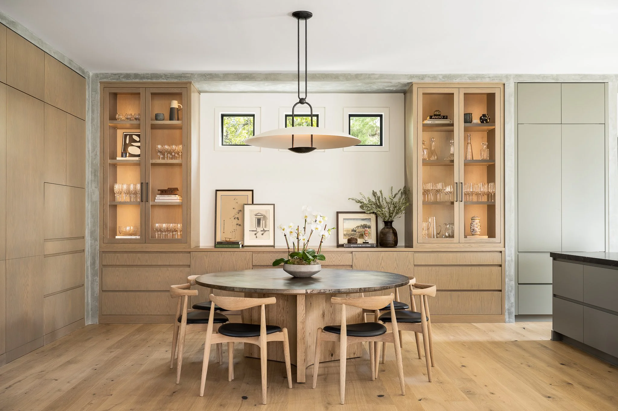 Modern dining room with a round wooden table, six chairs with black cushions, built-in wooden cabinets with glass doors displaying glassware, framed art, potted plants, and a large pendant light overhead. Small windows at the top provide natural ligh