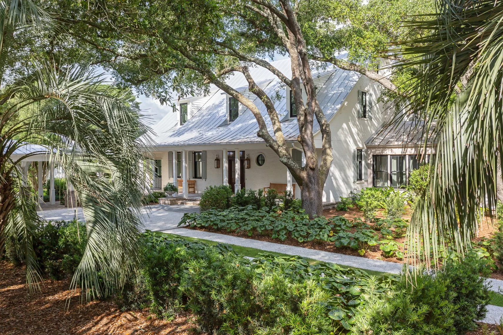 White house surrounded by lush greenery, trees, and plants, with a small front porch and a concrete walkway leading to the entrance.