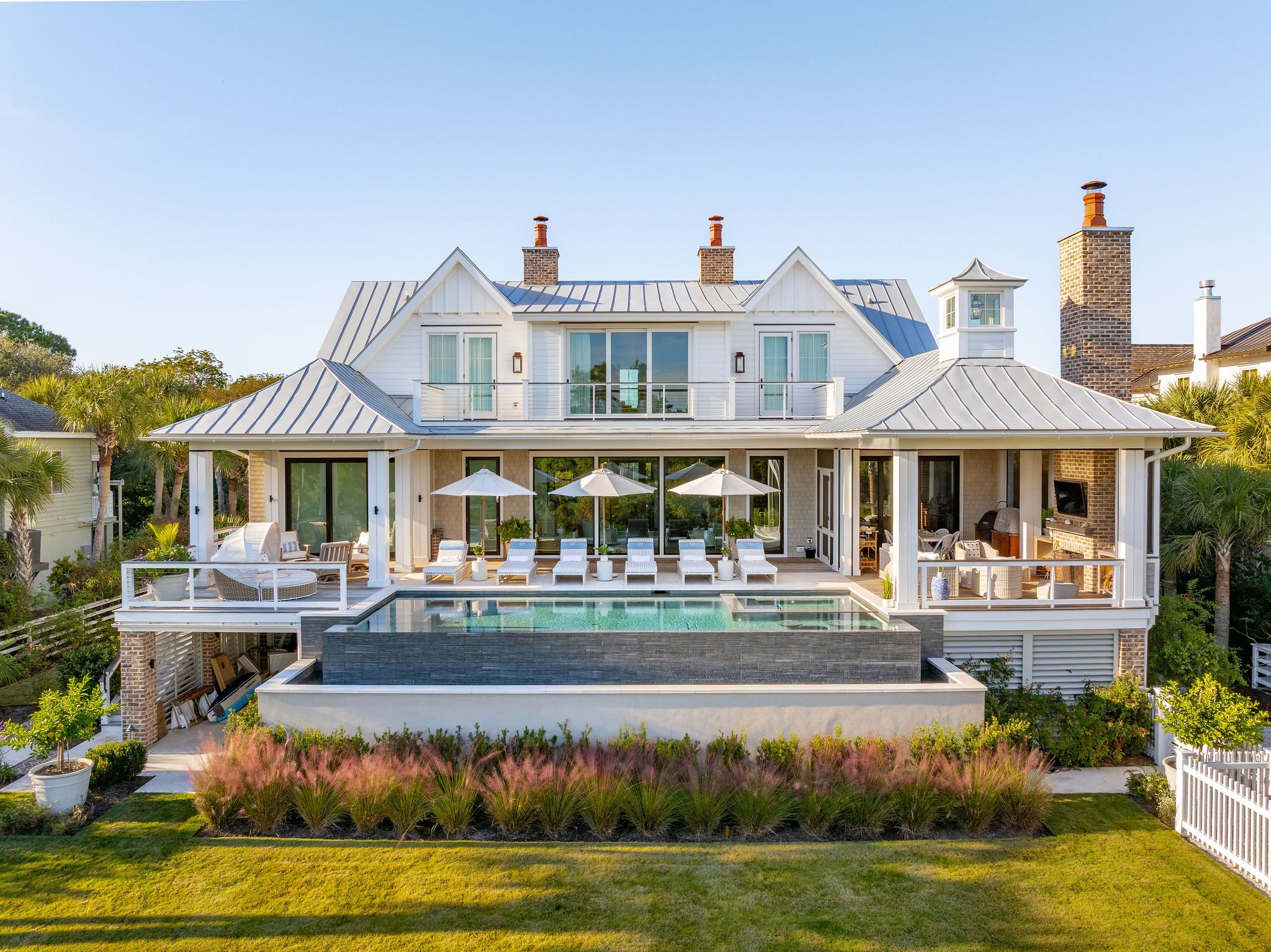 A large modern house with a white exterior, metal roof, and multiple chimneys. It has a pool area with lounge chairs, umbrellas, and outdoor seating. Surrounding greenery includes palm trees and flowering plants.