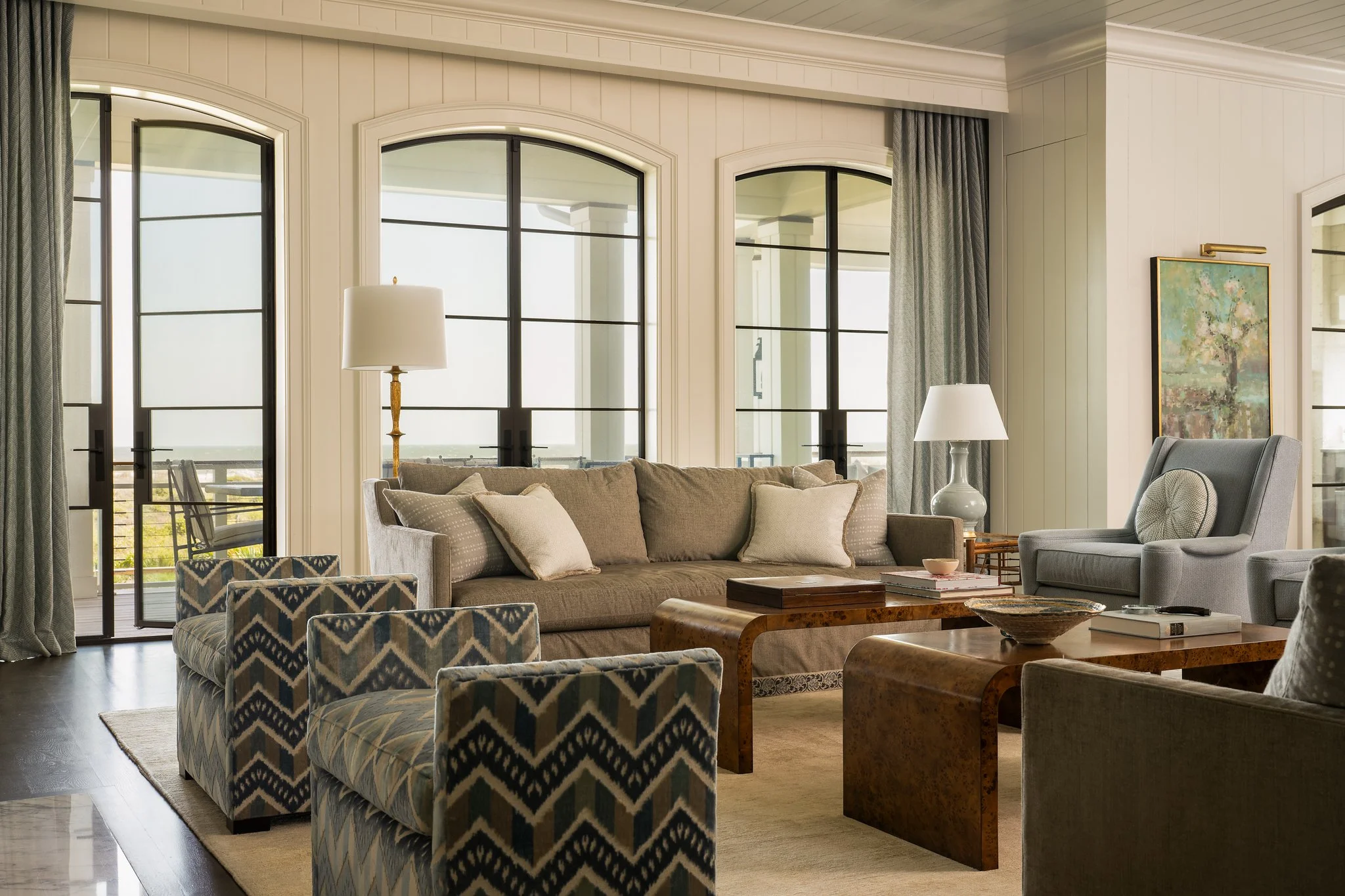 Living room with beige and gray furniture, large windows with black frames, and a balcony outside. Decor includes patterned armchairs, sofas with pillows, a wooden coffee table, and lamps, with curtains on the windows and a framed painting on the wal
