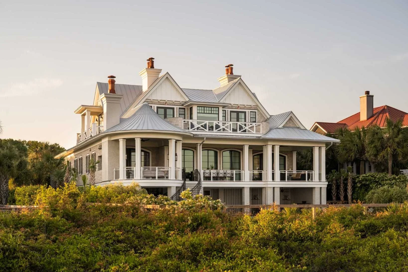 A large, multi-story house with a white exterior, metal roof, and multiple chimneys, surrounded by greenery and trees.