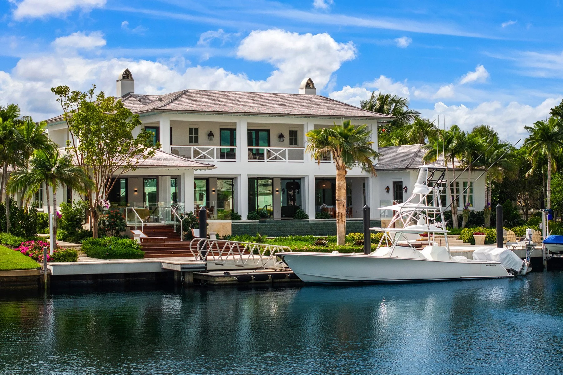 Luxury waterfront house with balconies, glass doors, lush gardens, palm trees, and boats docked at the marina, under a partly cloudy sky.