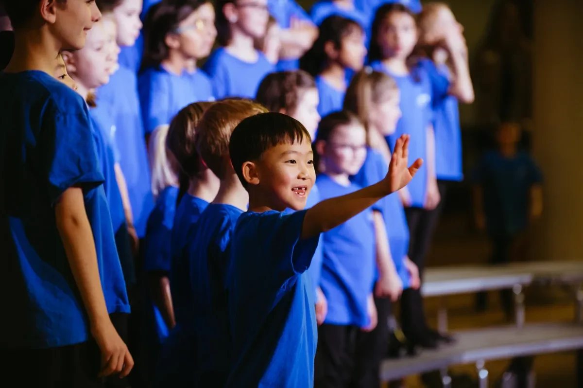Young boy in a blue choir shirt waves from the risers at a children's choir concert