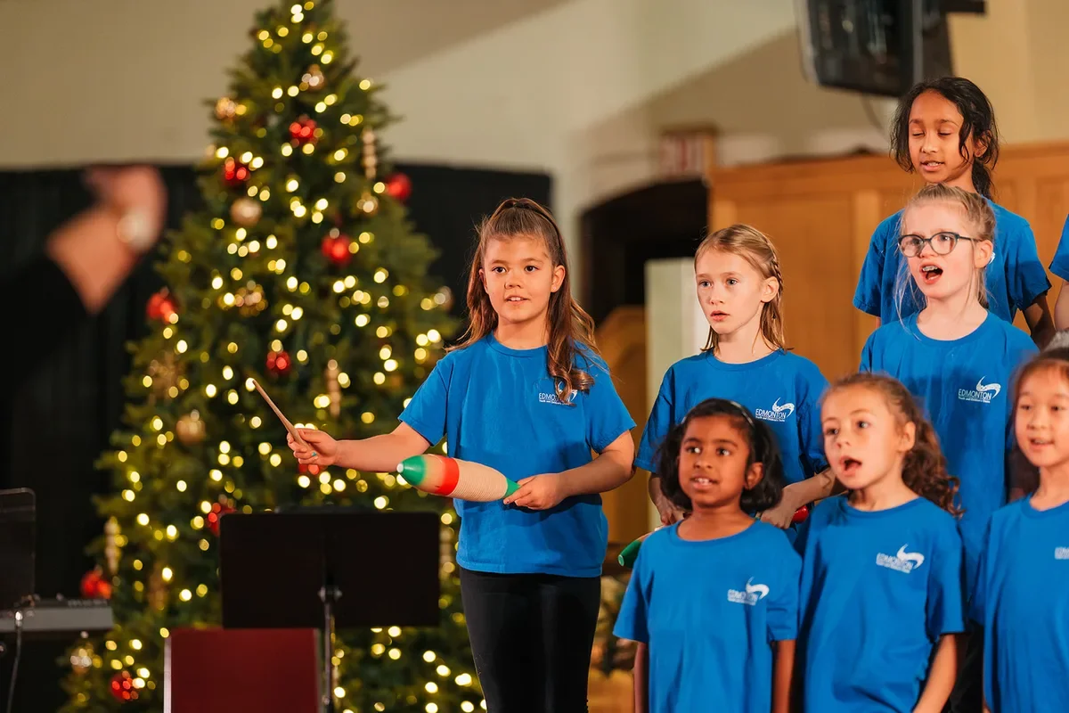 A girl plays a guiro while her fellow choir members sing beside a lit Christmas tree