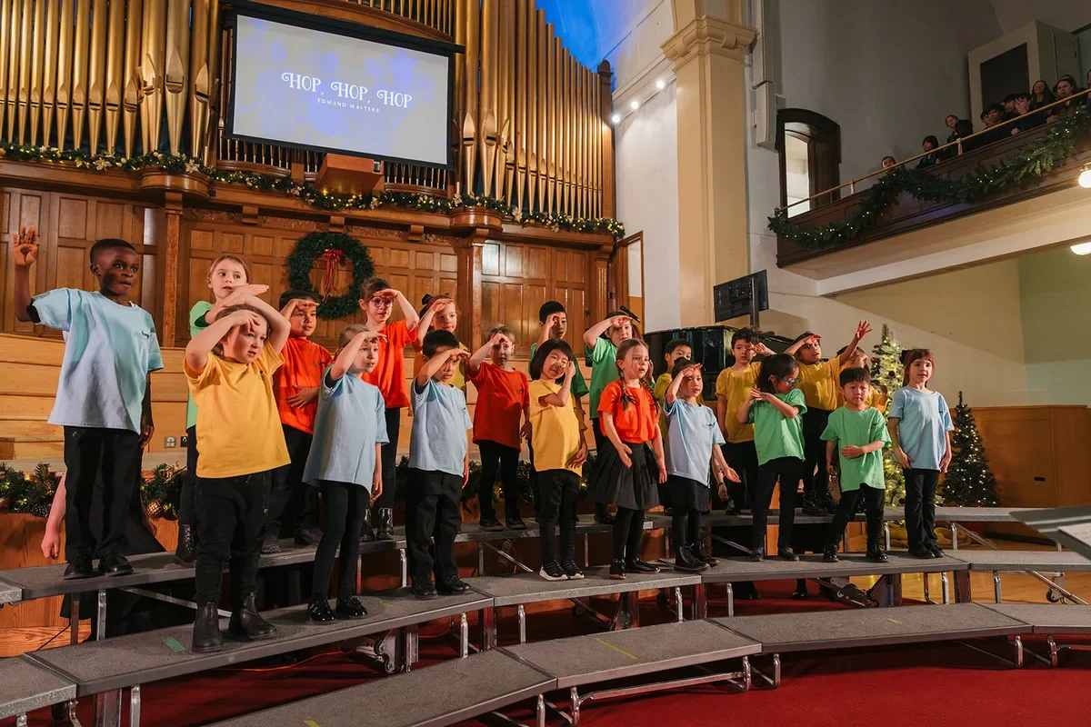 Young singers in colourful T-shirts do hand actions to a song on stage at a holiday concert
