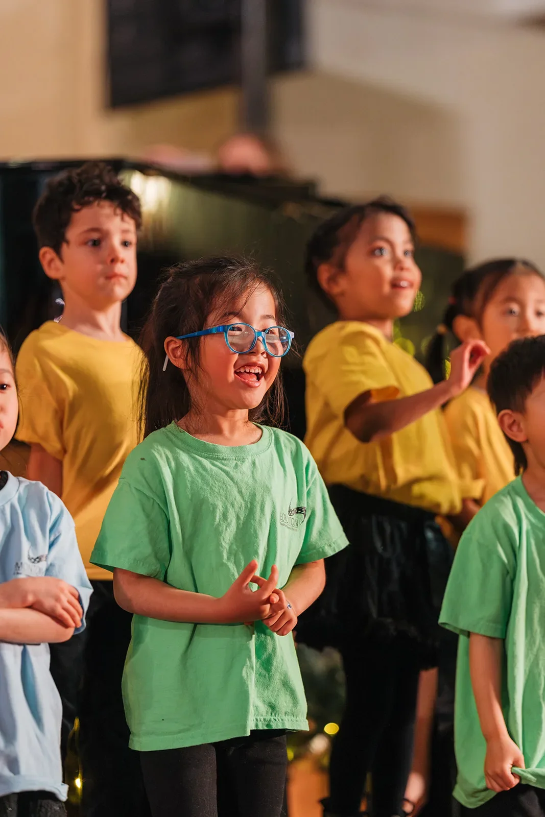 A young singer in blue glasses and a green T-shirt smiles while performing with her choir