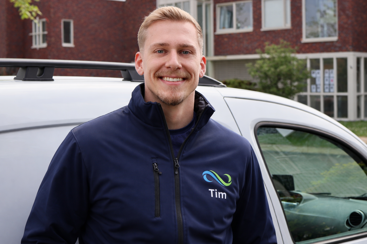 Vrouw met blond haar en een blauwe jas met de naam Tim, tevreden lachend naast een witte bedrijfsauto met appartementen op de achtergrond.