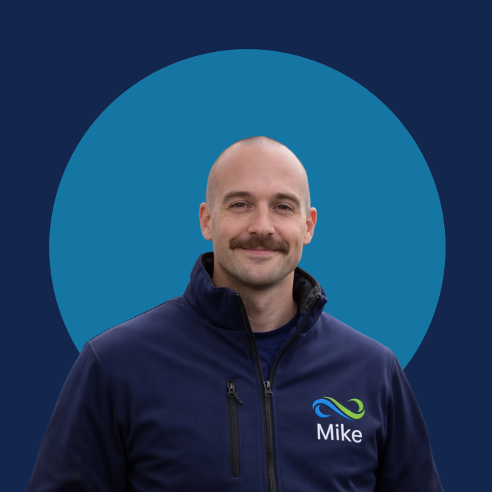 Een man met een bald head en snor, glimlachend, gekleed in een donkerblauwe jas met een logo en de naam 'Mike', tegen een achtergrond met een grote blauwe cirkel op een donkerblauwe achtergrond.