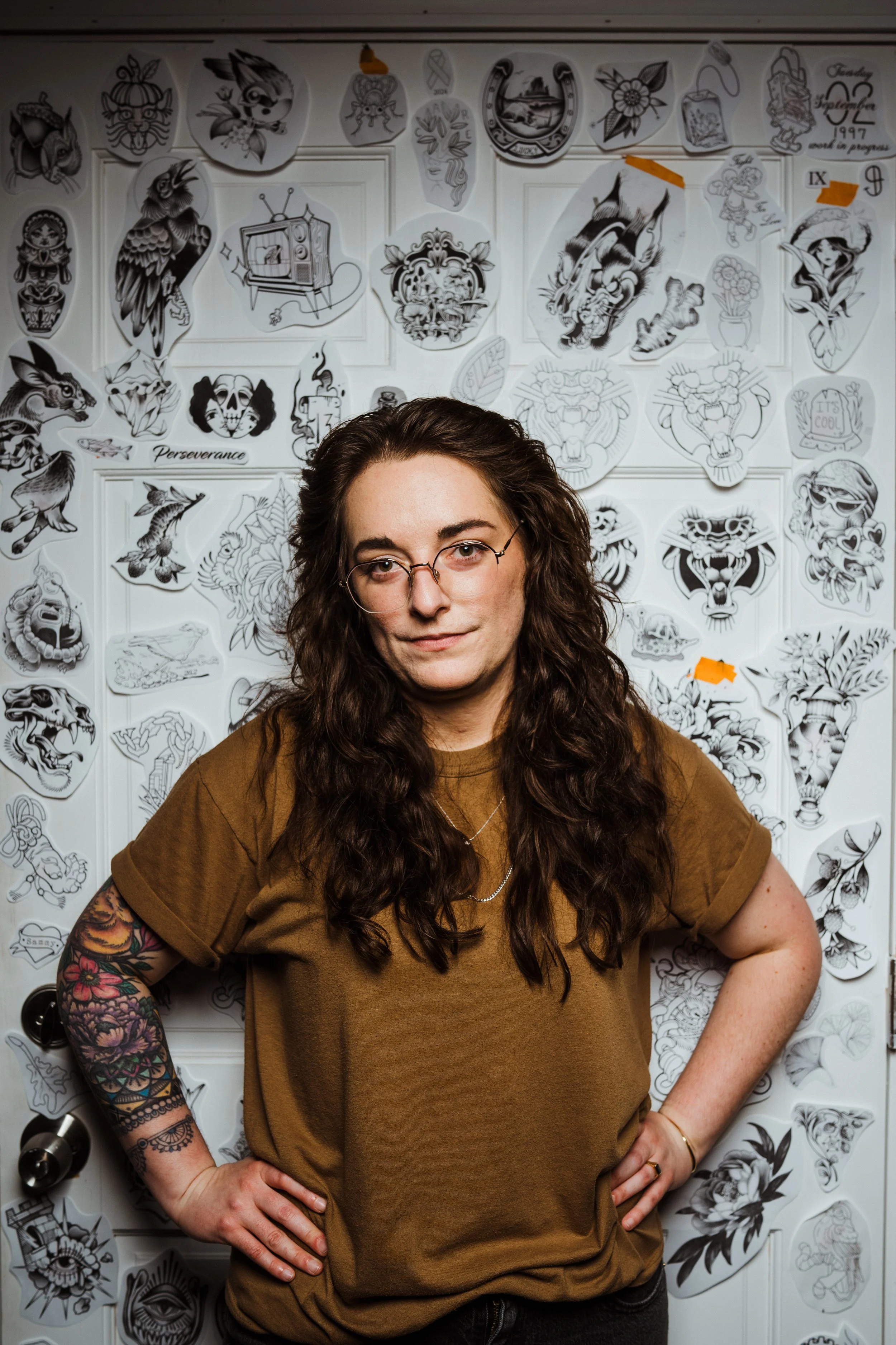 Woman with long, curly dark hair, glasses, wearing a brown T-shirt, standing in front of a wall decorated with various black and white tattoo-style illustrations.