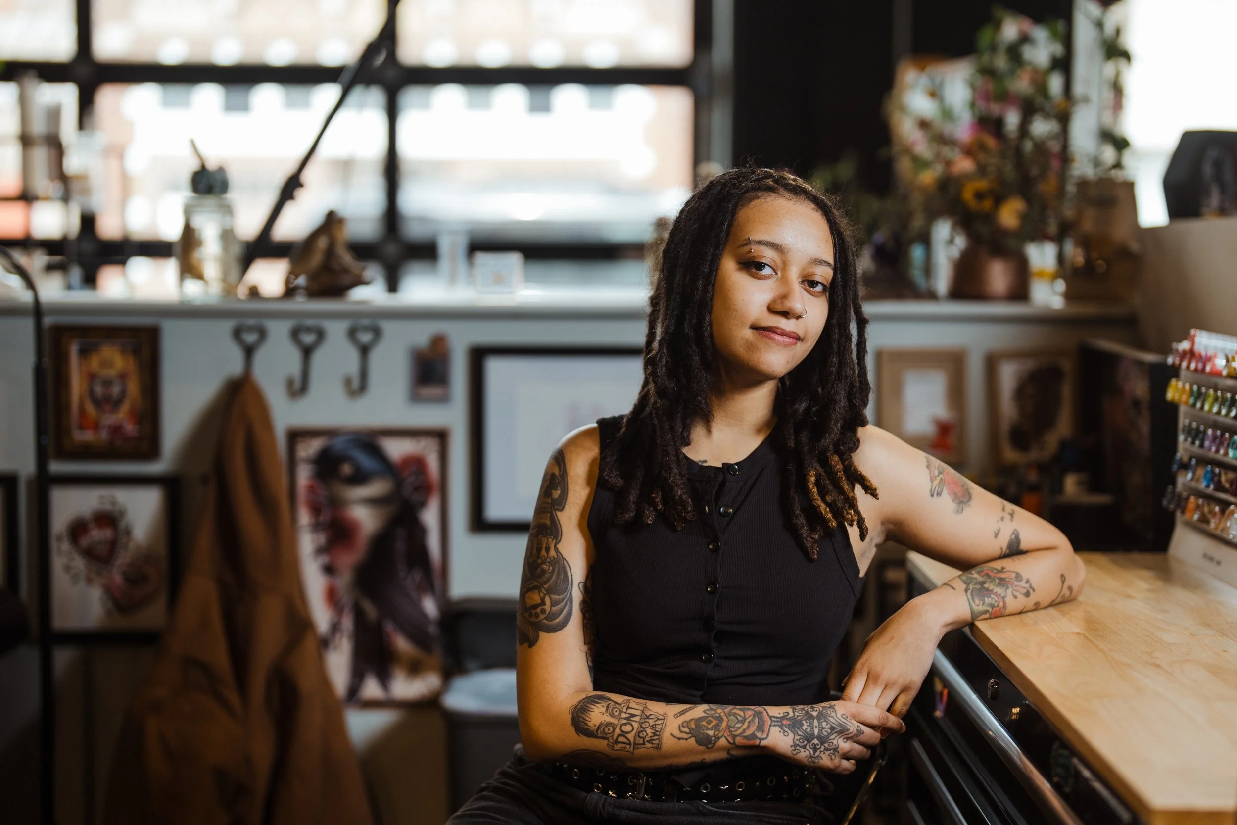 A young woman with tattoos on her arms and dreadlocks sitting at a bar or counter inside a tattoo studio, with framed art, keys, and flowers in the background.