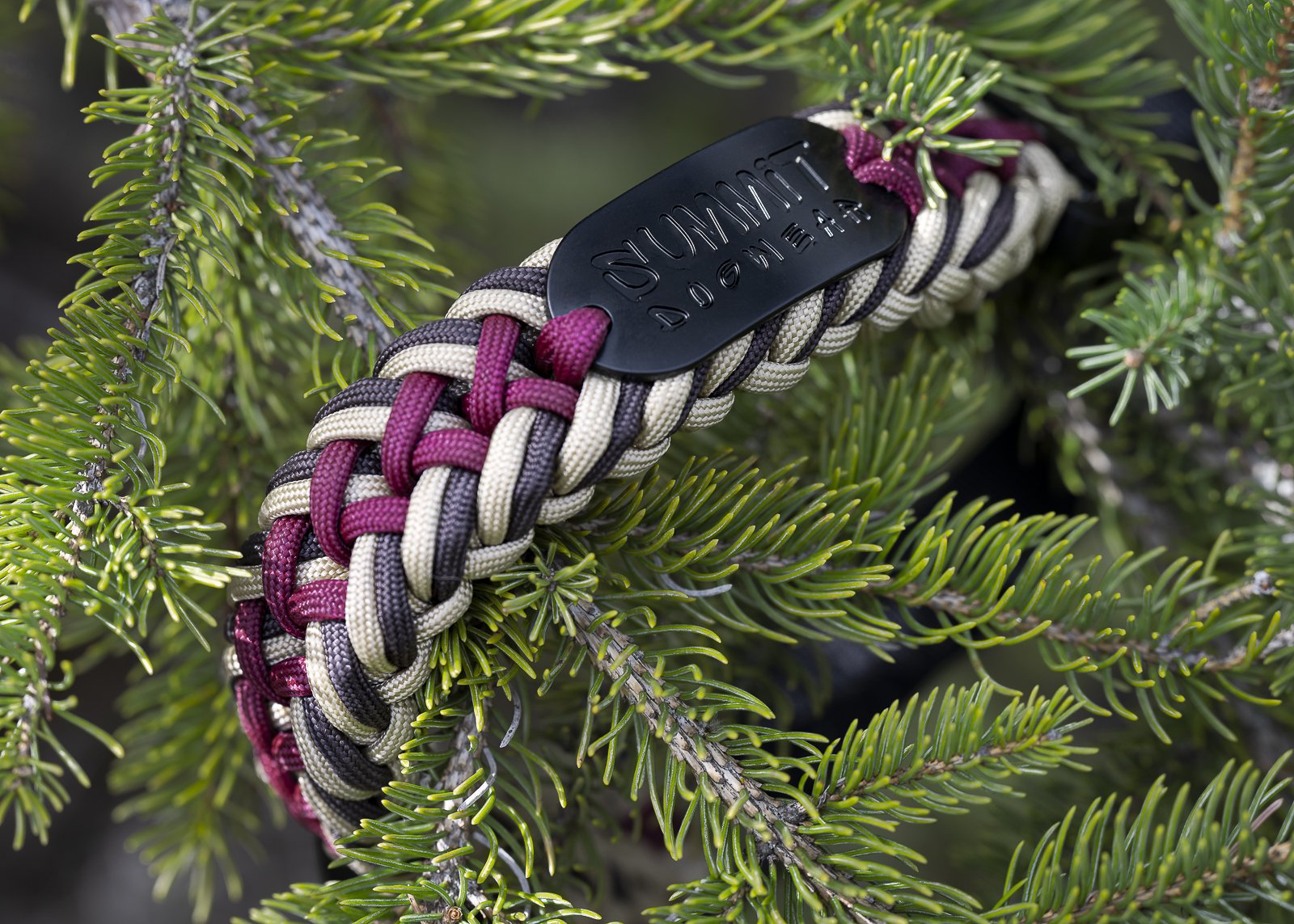 A paracord bracelet with a black rubber clasp engraved with the word 'NOMAD' on a green pine tree branch.