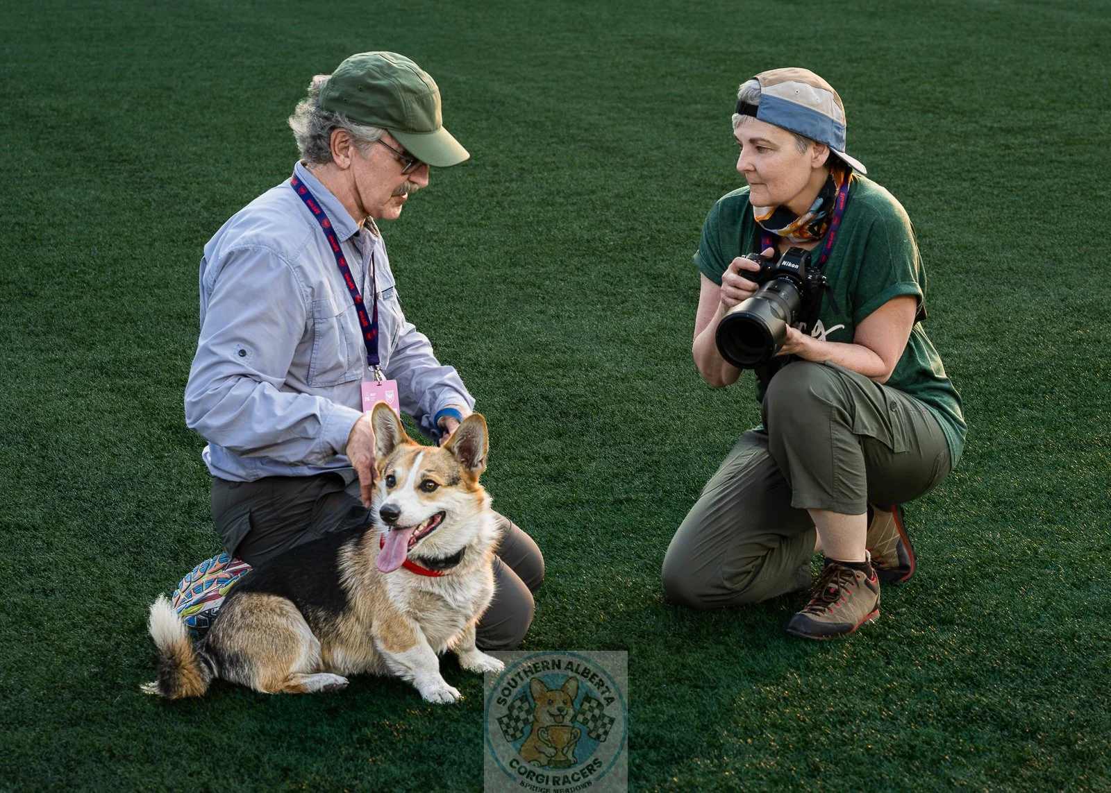 Two people kneeling on a grassy field with a corgi dog. One person is holding the dog and the other is taking a photo of the dog, which has its tongue out.