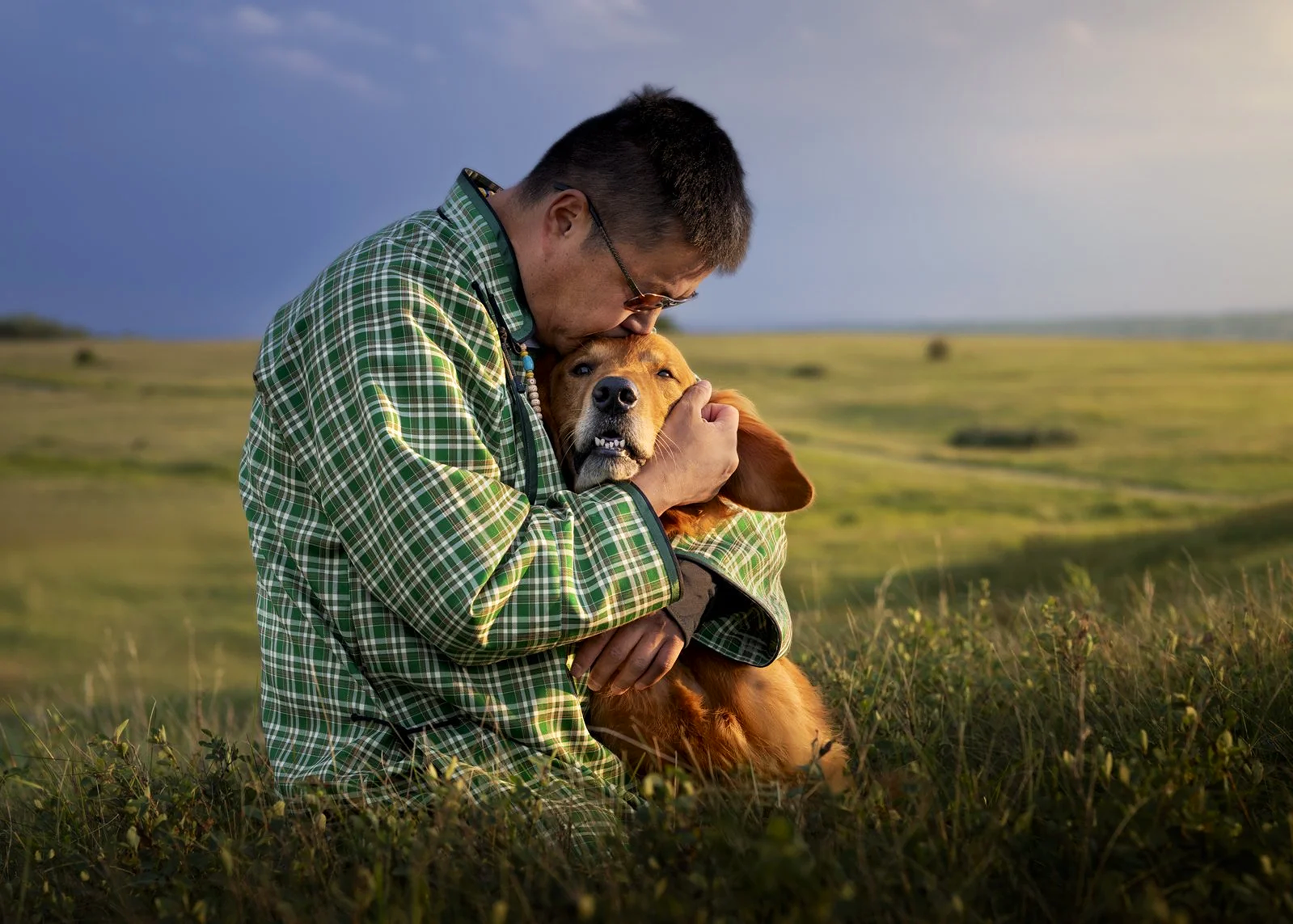 A man in a green plaid shirt embraces a golden retriever dog in a grassy field, with a landscape and cloudy sky in the background.