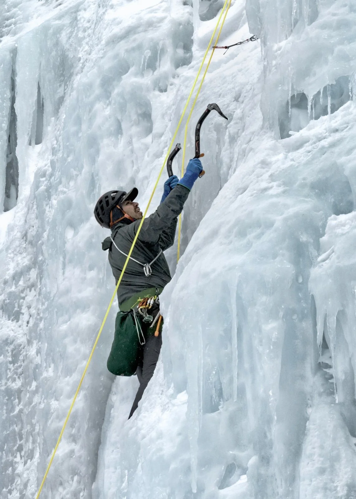 A person ice climbing on a frozen ice wall, holding two ice axes and secured with climbing gear, wearing a helmet, gloves, and a harness.