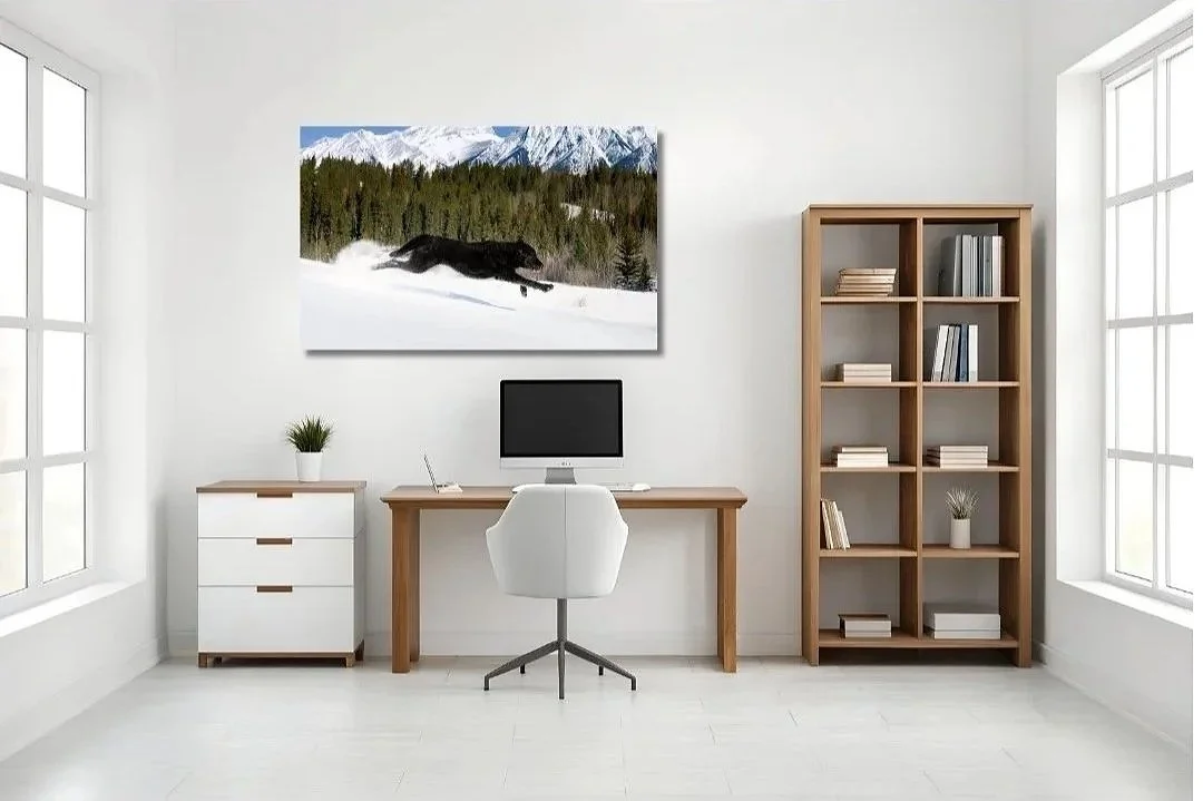 Minimalist home office with white walls, large windows, a wooden desk with a computer, a white office chair, a small white cabinet with a plant, a tall wooden bookshelf with books, and a landscape painting of snow, trees, and mountains hanging above the desk.