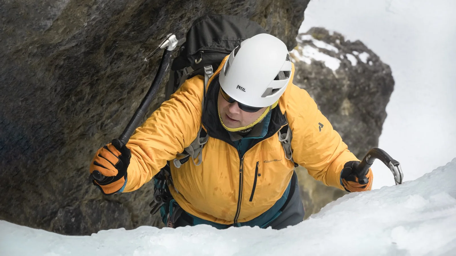 Male climber in a yellow jacket and helmet ascending a snowy slope with rock formations nearby.