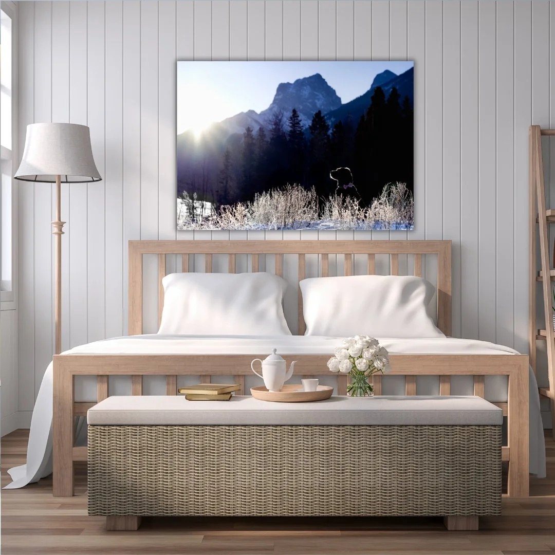 A bedroom with a wooden bed, white bedding, and a bench at the foot of the bed with a teapot, cup, books, and a vase of white flowers. There is a painting of mountains and trees above the bed, and a floor lamp to the left. white paneled walls and a window are visible.