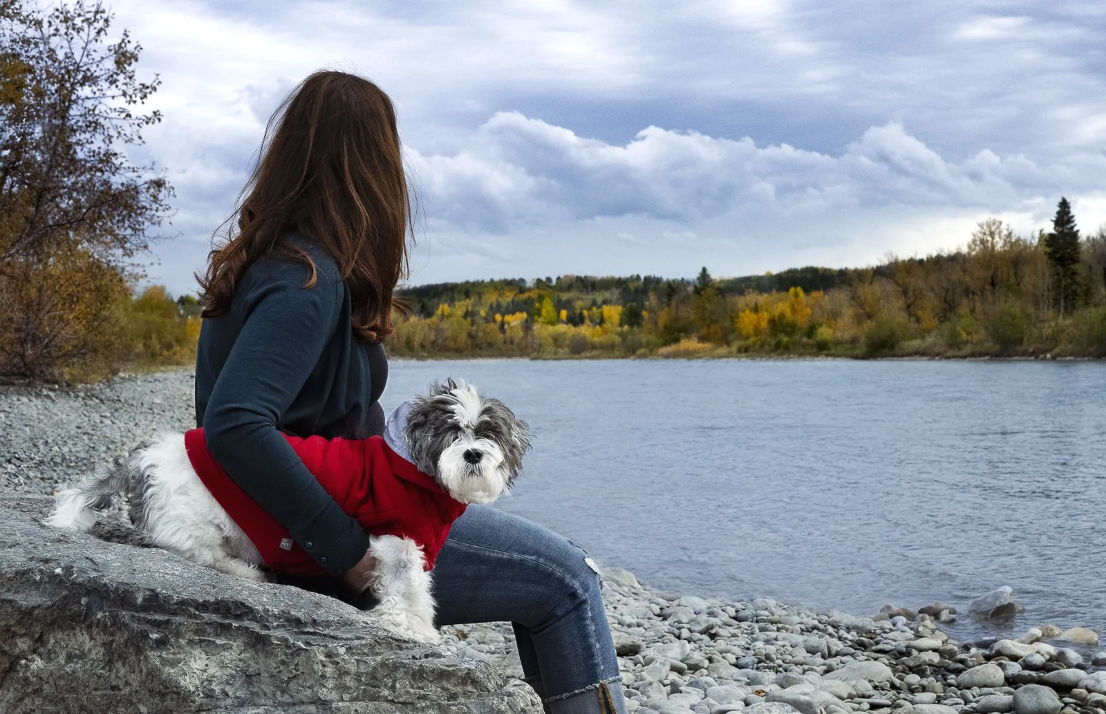 A woman with brown hair sitting on a rock by a river, holding a small black and white dog in a red jacket, with trees and cloudy sky in the background.