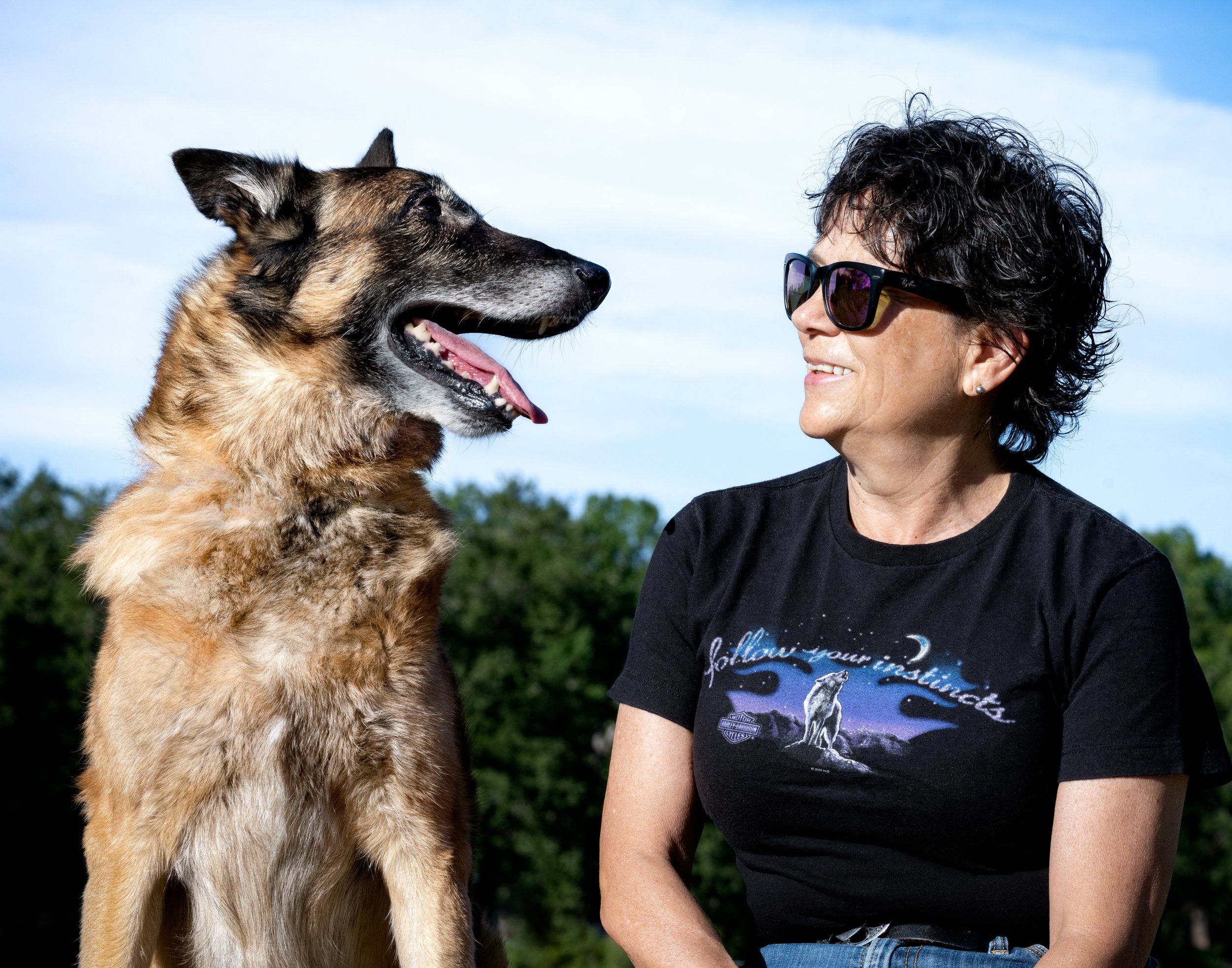 A woman with short curly black hair and sunglasses smiling at a large belgian malinois dog outdoors against a blue sky and green trees.