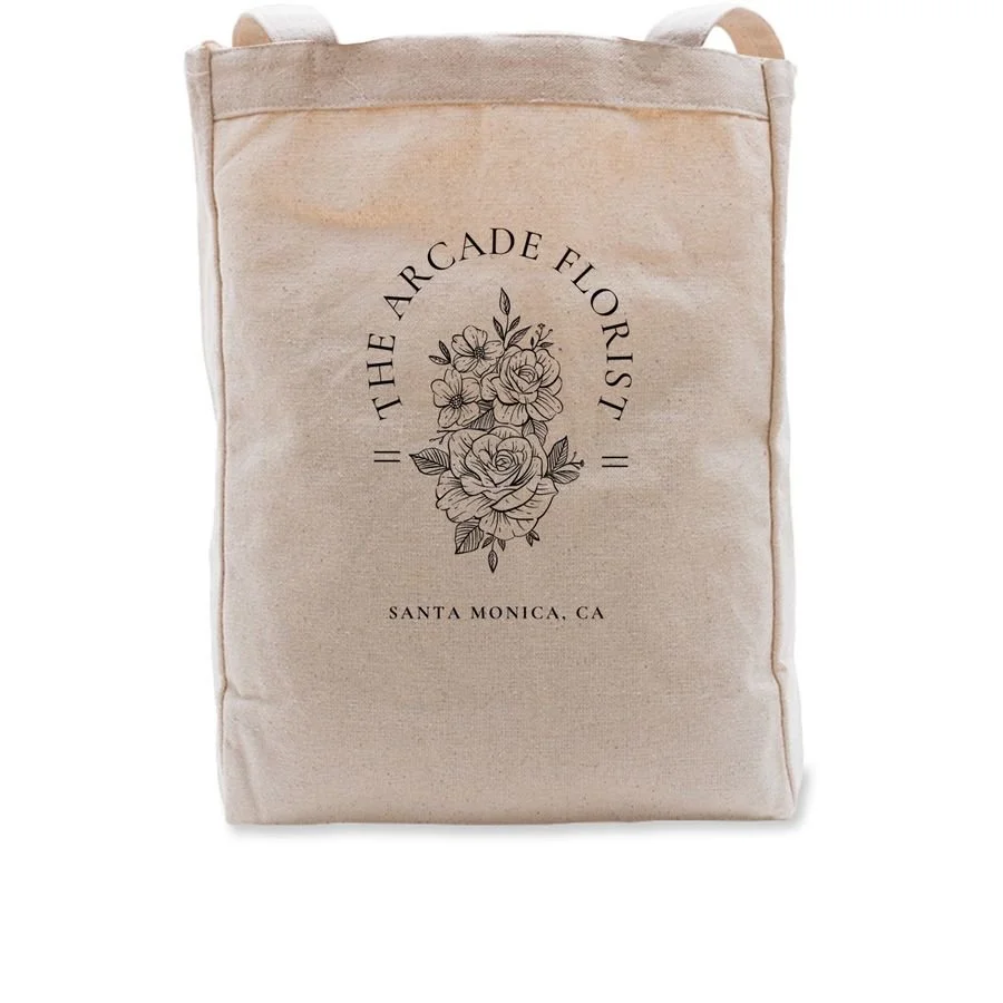 Beige tote bag with black floral design and text reading "The Arcade Florist, Santa Monica, CA".