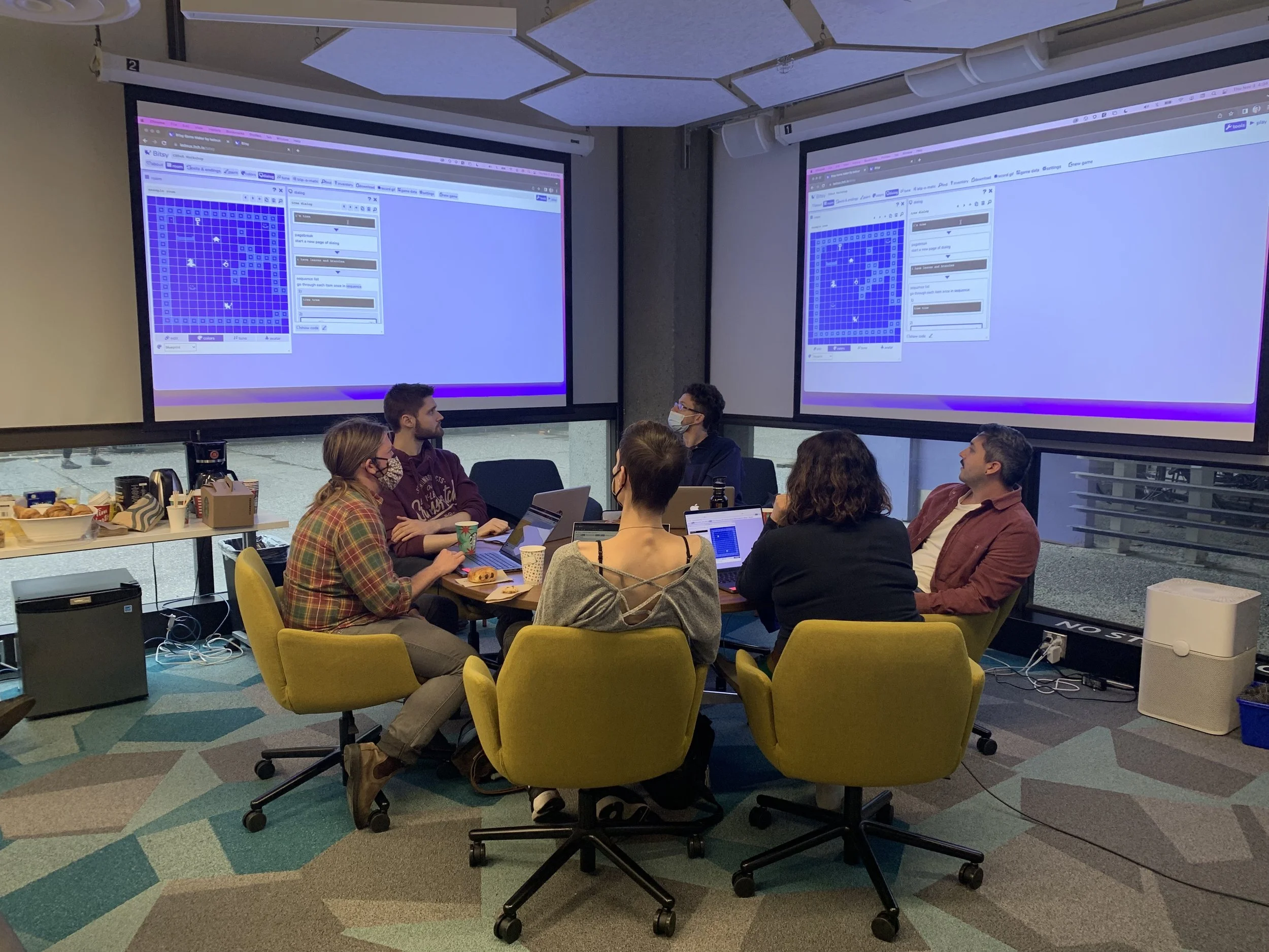 Six people sitting around a circular table, on yellow neon swivel chairs. There is a blue projection screen behind them, and a geometric patterned carpet underneath.
