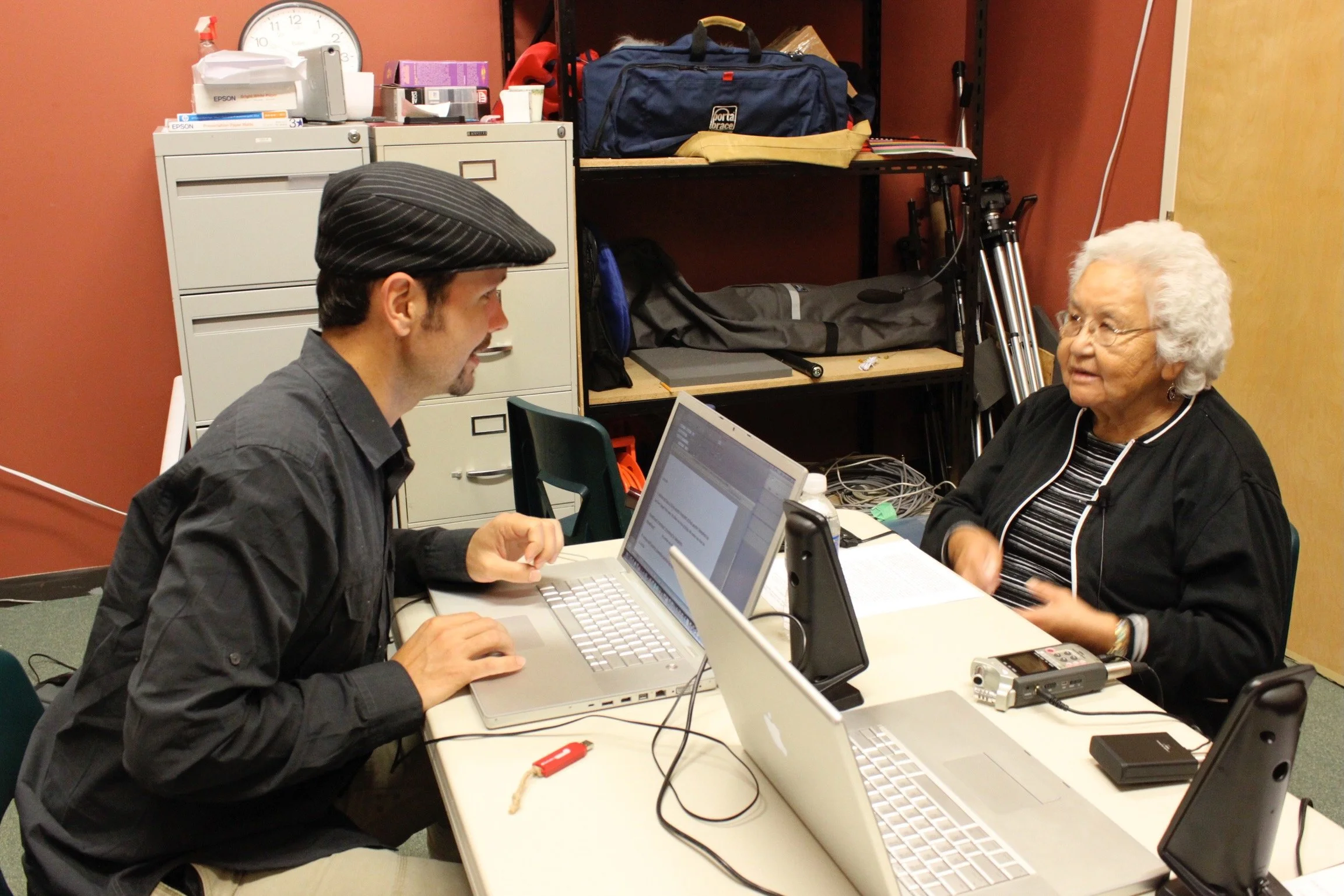 Two people facing one another on opposite sides of a white desk with two laptops between them. Laptops facing opposite directions on the table. The person on the left is leaning forward to the person on the right.