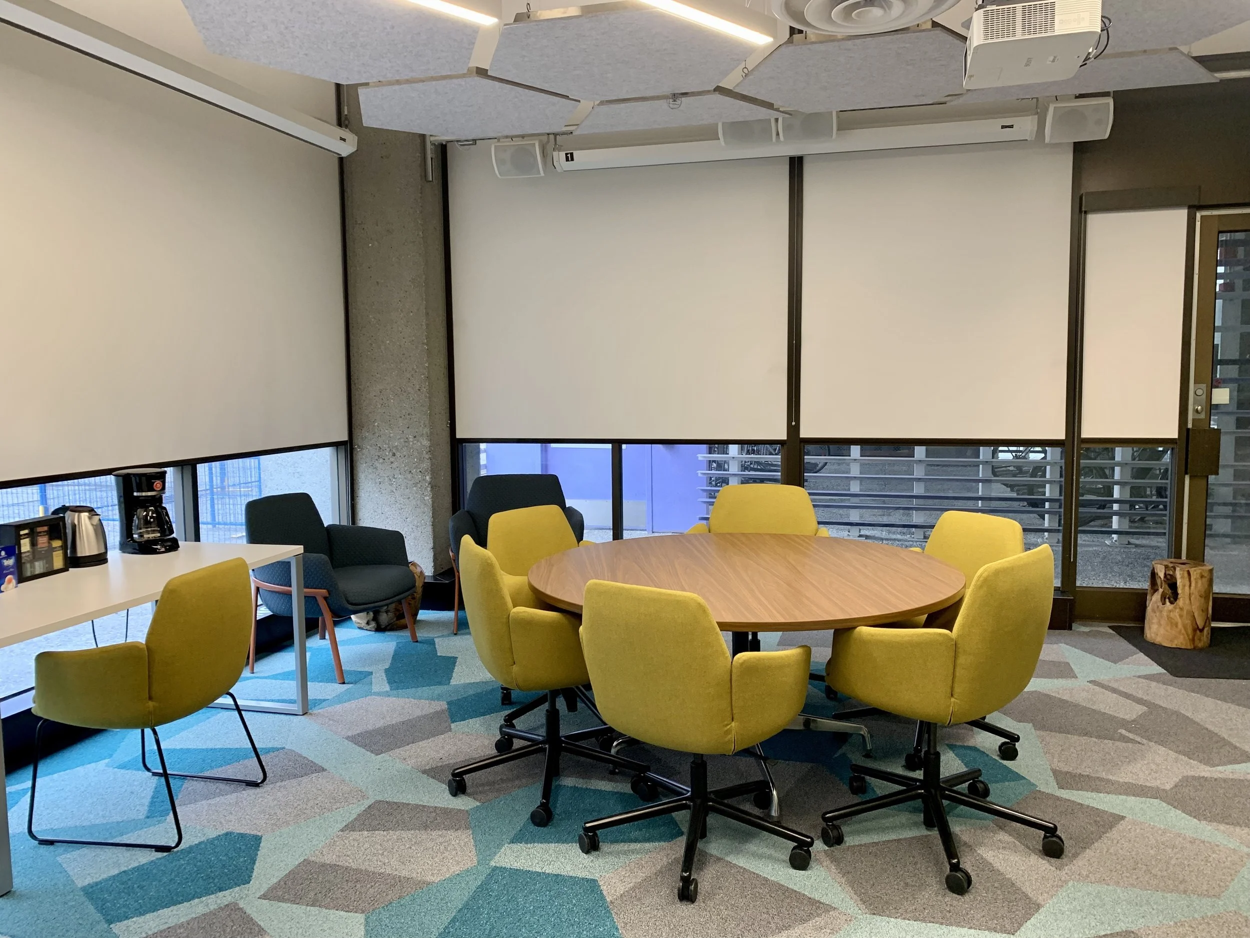 Image: Modern, brightly lit, collaborative office setting at CEDaR, with a round wooden table surrounded by six yellow swivel chairs.