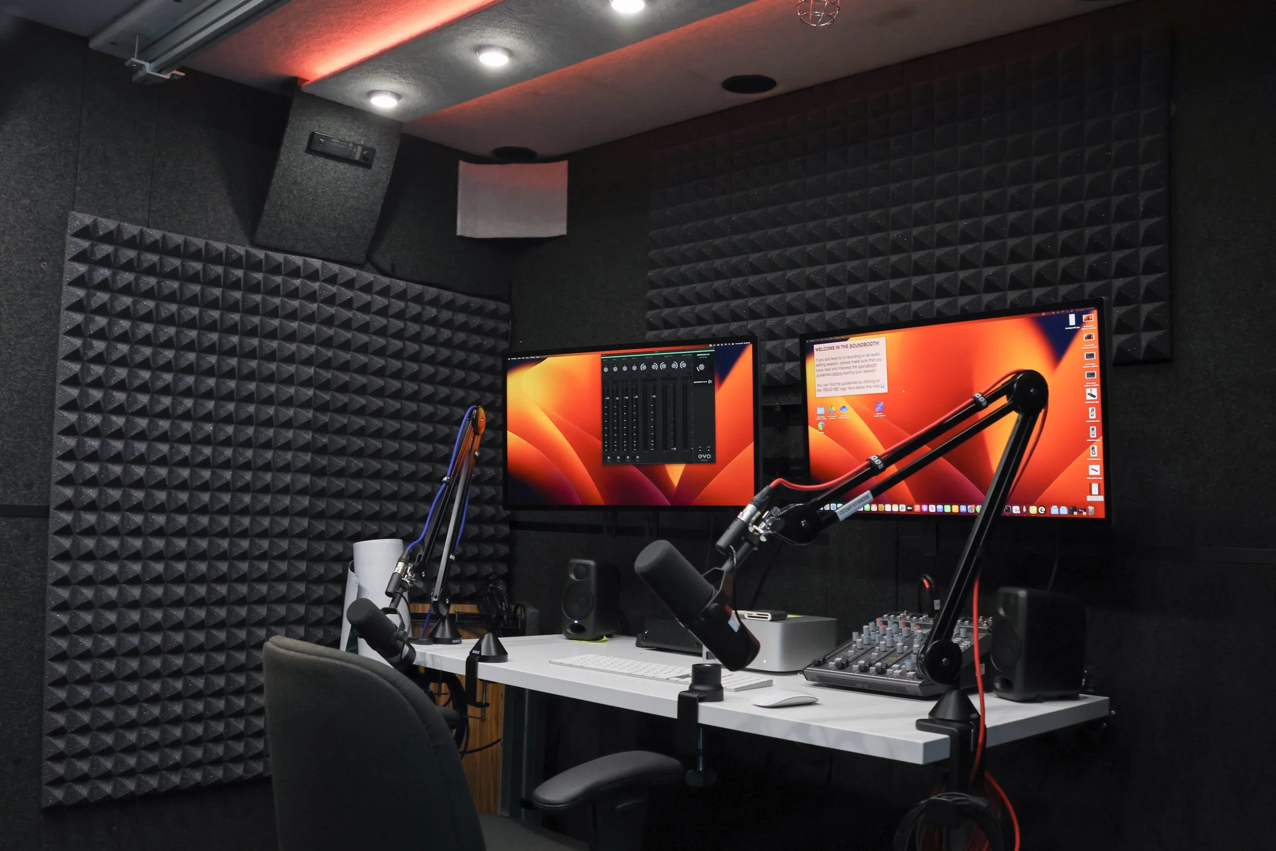 A soundbooth at CEDaR Space with padded black walls and a white desk in the center. There are mics attached to the desk, facing towards a grey chair in-front of the desk. There are two large computer monitors with glowing orange screens opposite