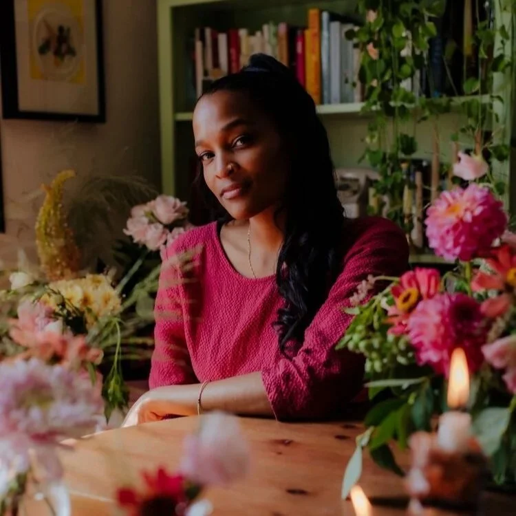 A woman with long dark hair, wearing a maroon sweater, sitting at a wooden table surrounded by colorful flowers and a lit candle, with a bookshelf and green plants in the background.