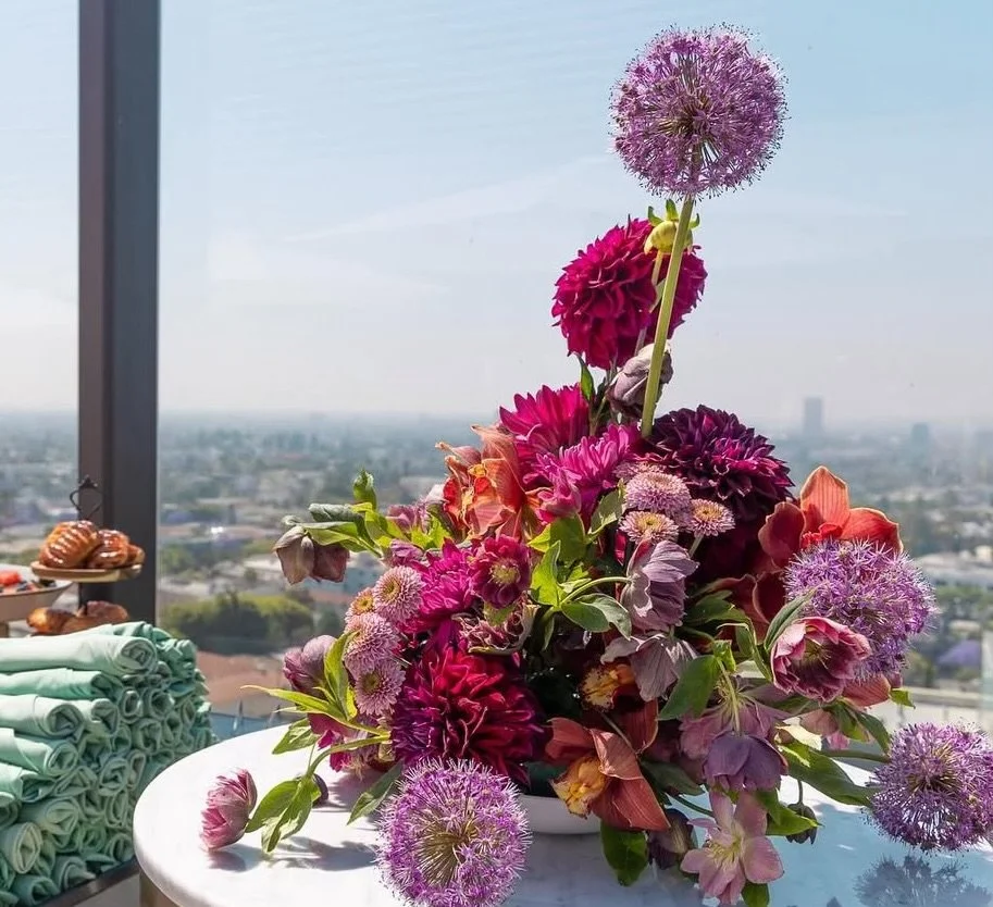 A colorful flower arrangement with pink, purple, and red flowers on a white table in front of a large window overlooking a cityscape.