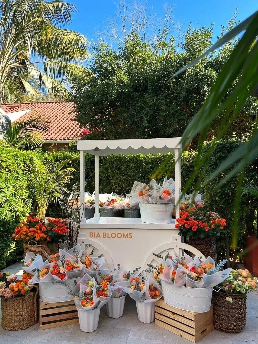 Flower cart with bouquets of orange, pink, and yellow flowers, surrounded by baskets and wooden crates, set outdoors against lush greenery and a blue sky.