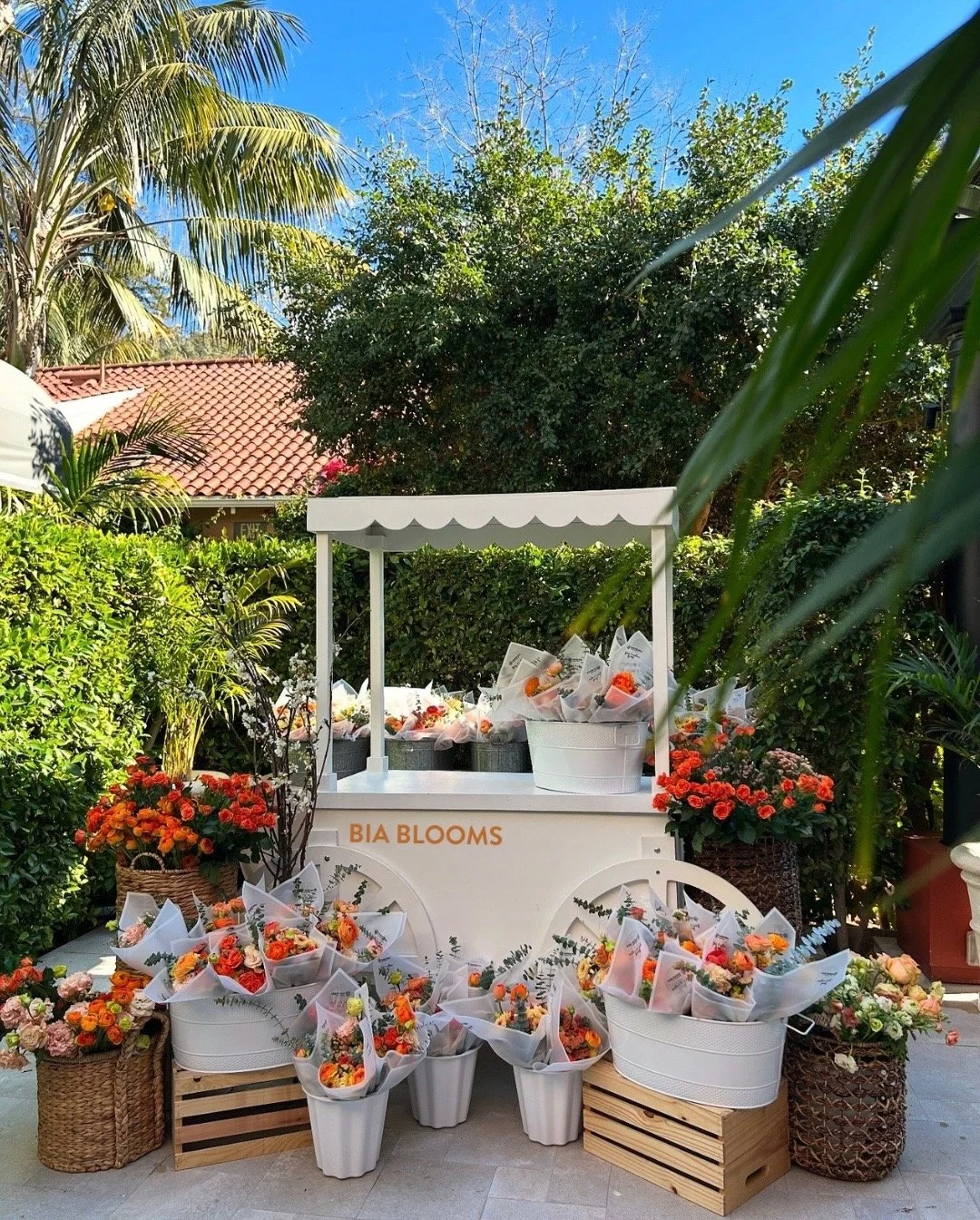 A flower stand called "BIA BLOOMS" with various flower bouquets displayed on tables, baskets, and crates outdoors, surrounded by greenery and trees, under a blue sky.