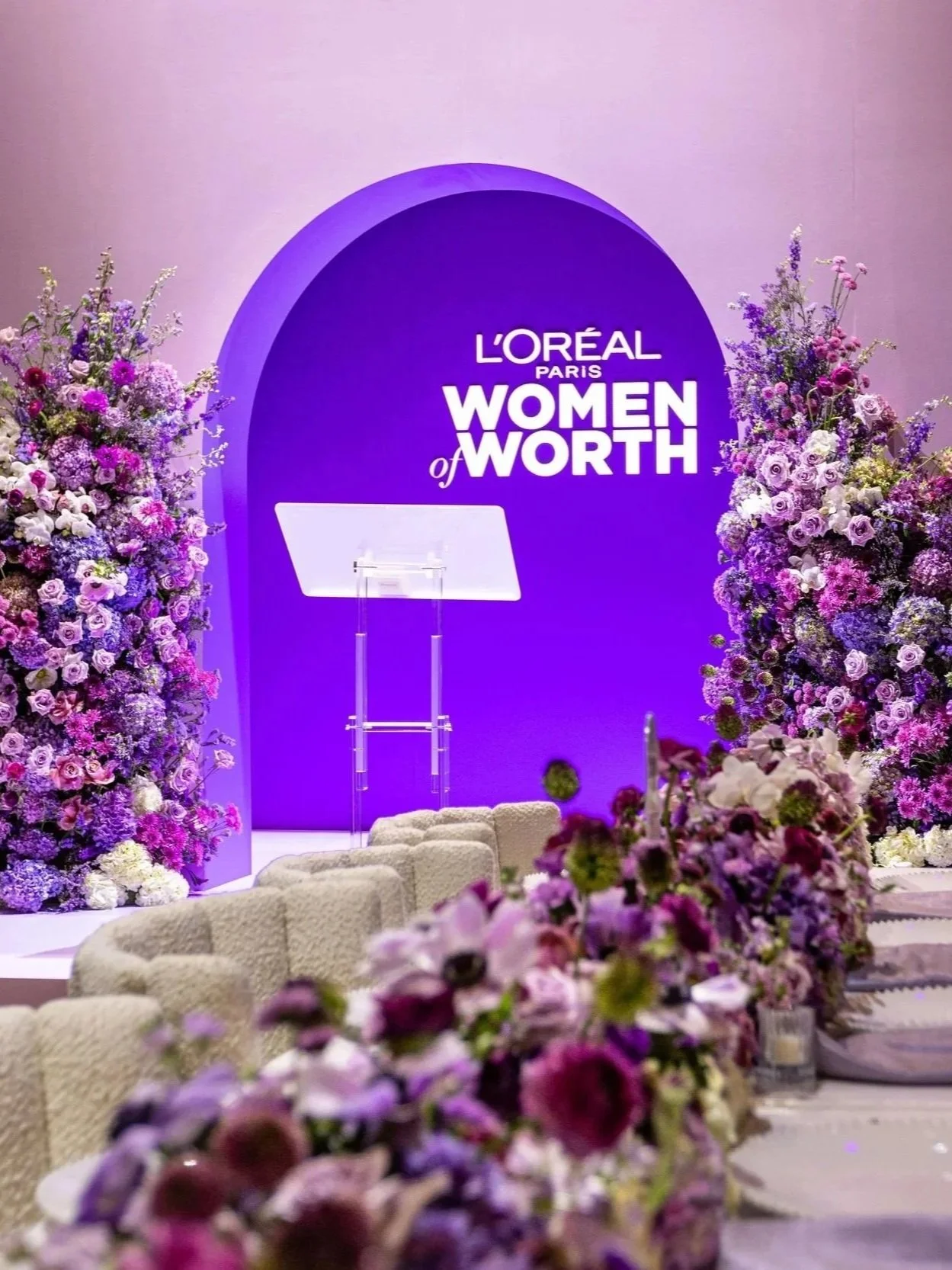 Stage decorated with purple flowers and large purple backdrop with 'L'Oreal Paris Women of Worth' logo, set up for an event or ceremony.