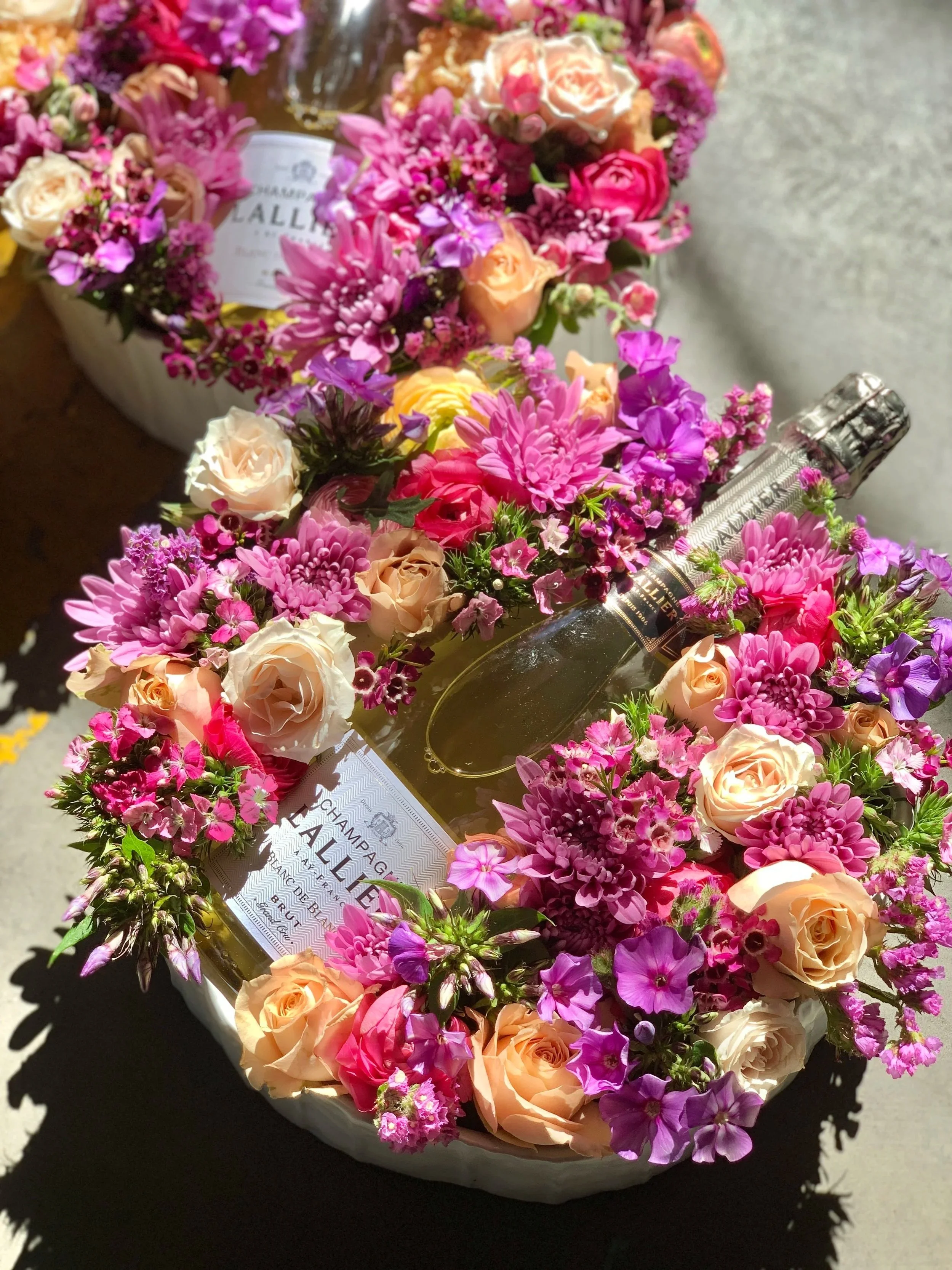 A collection of pink and peach-colored flowers, including roses and asters, arranged in a decorative container. Some flowers have labels indicating they are from 'Champagne Lallier,' a champagne brand, suggesting that the flowers may be a gift or part of a celebration.