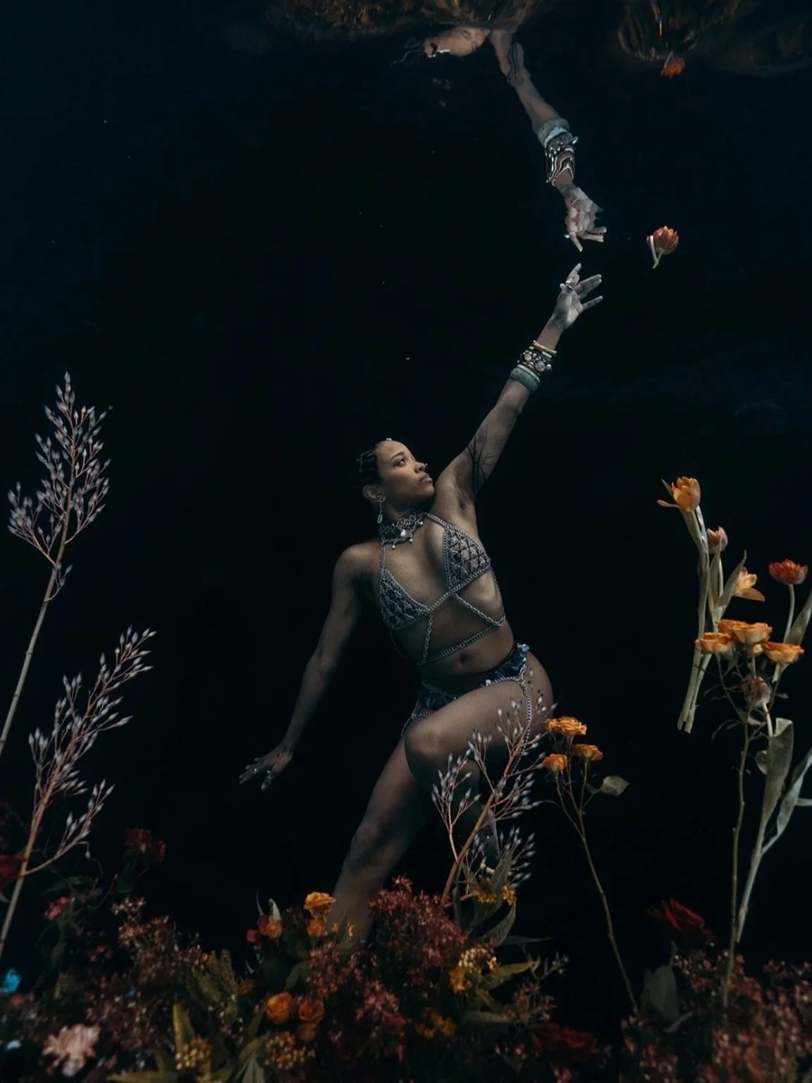 A woman wearing a revealing, bejeweled outfit with jewelry reaches upward with her right arm, surrounded by tall flowers and plants, in a dimly lit setting.