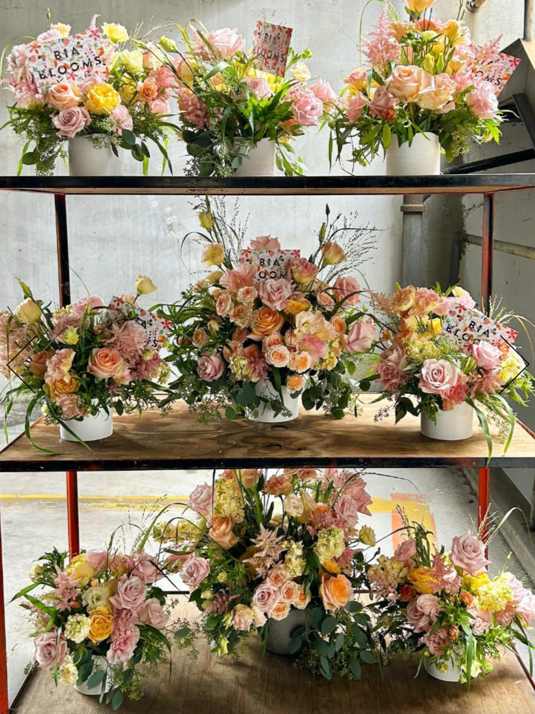 Multiple floral arrangements with pink, yellow, and white flowers, including roses, displayed on a multi-tiered shelf.