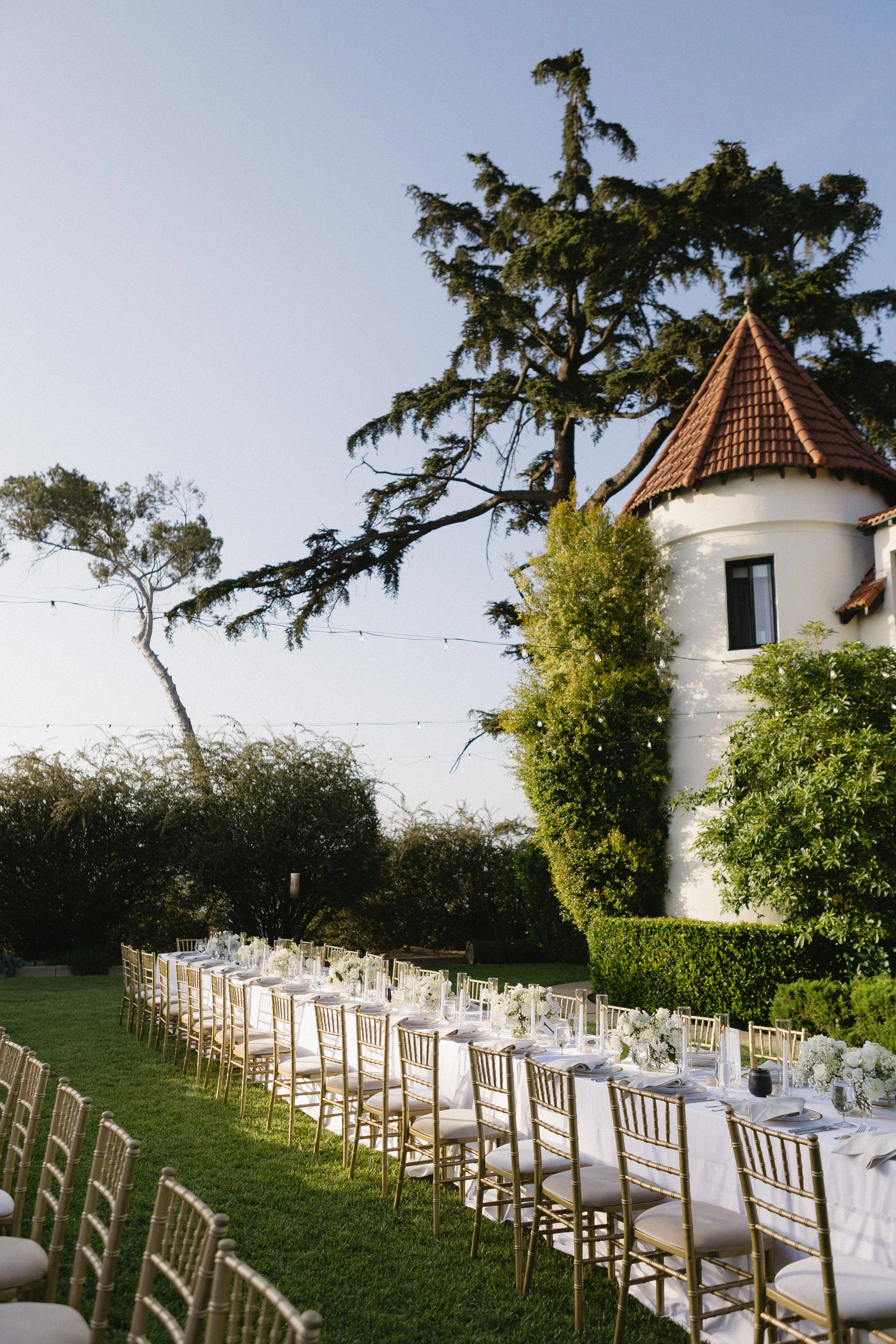 Outdoor wedding reception setup with a long white table, gold chairs, floral centerpieces, and a historic building with a tower and tree in the background.
