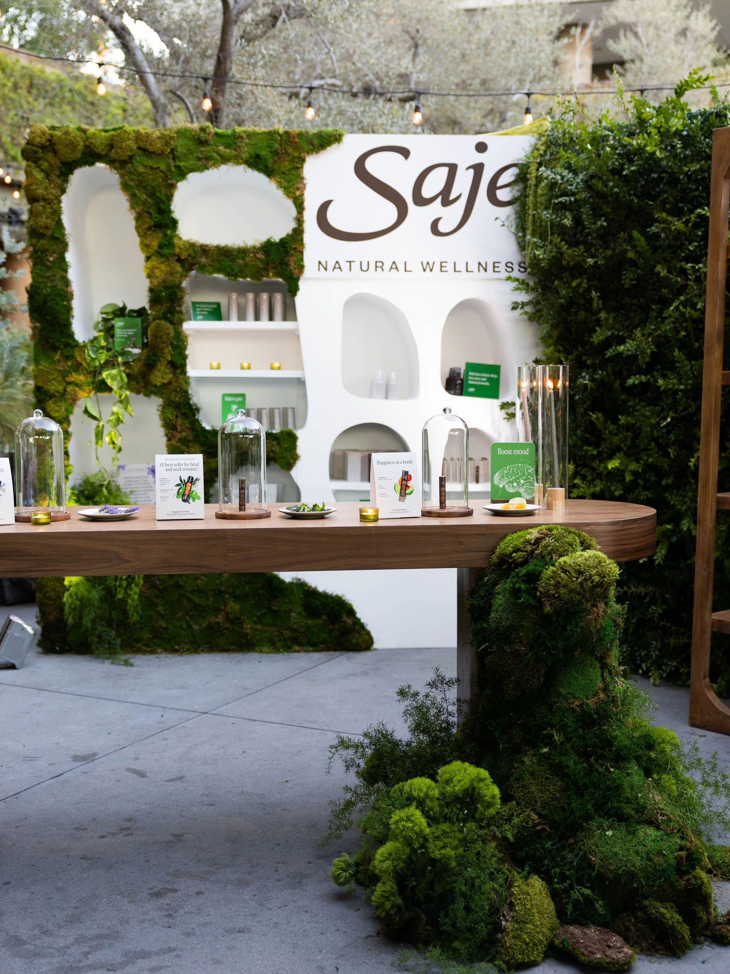 An outdoor display with a wooden table, glass displays, and small plants, in front of a white wall with the word 'Saje' and 'Natural Wellness.' There are greenery, moss, and trees in the background.