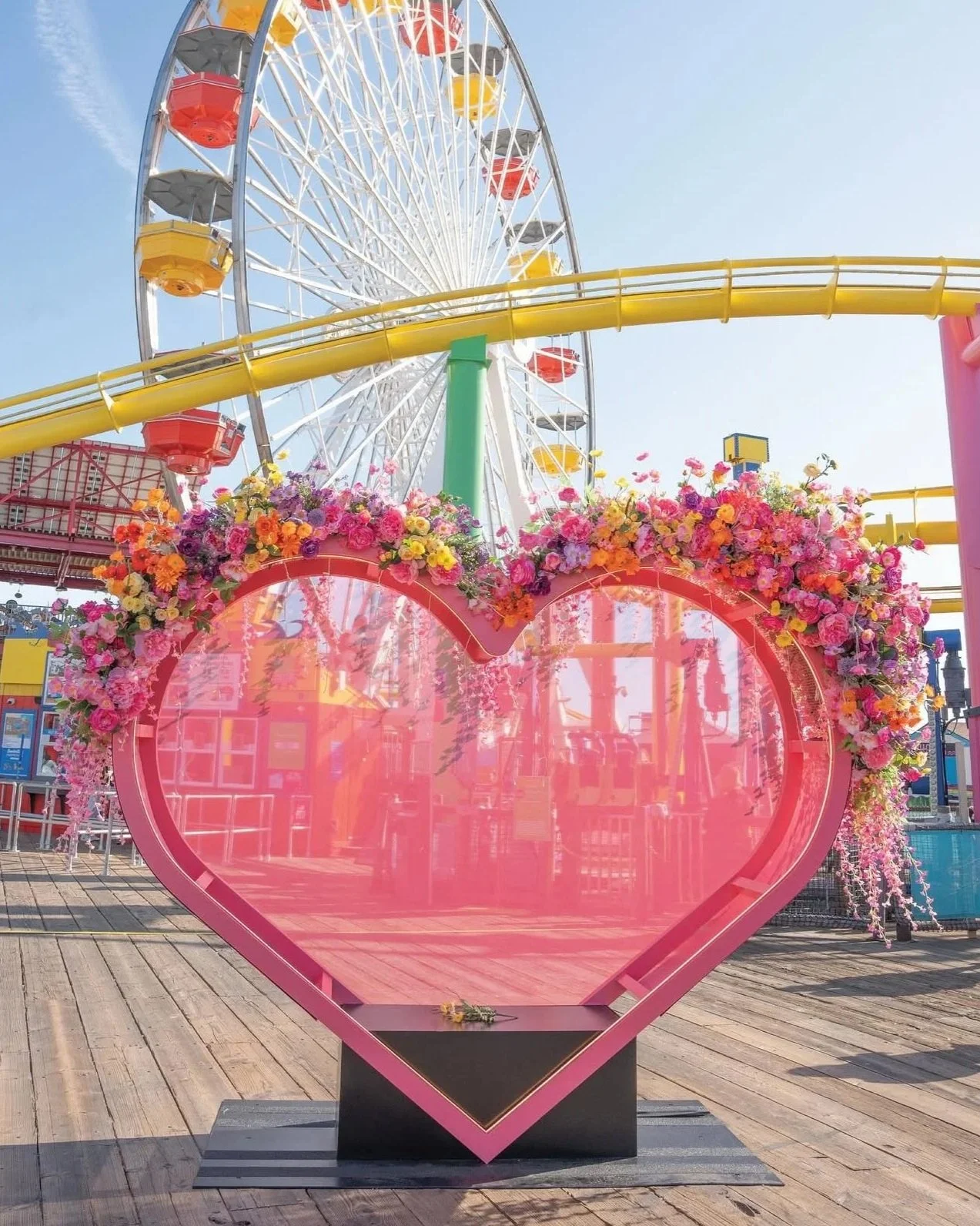 Colorful Valentine's Day display with a large pink heart-shaped frame decorated with pink, orange, and yellow flowers, and a Ferris wheel in the background at an amusement park.