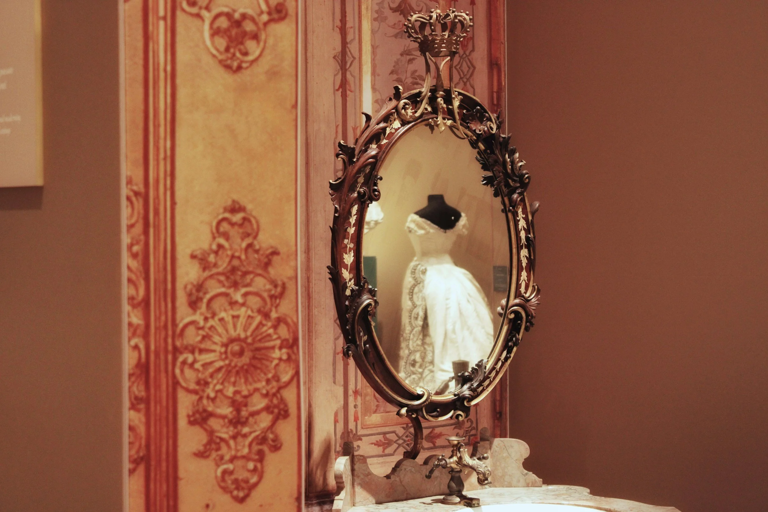 An ornate, vintage gold oval mirror on a decorative pink wall reflecting a ghostly white figure, used when curating photos for historical fiction authors