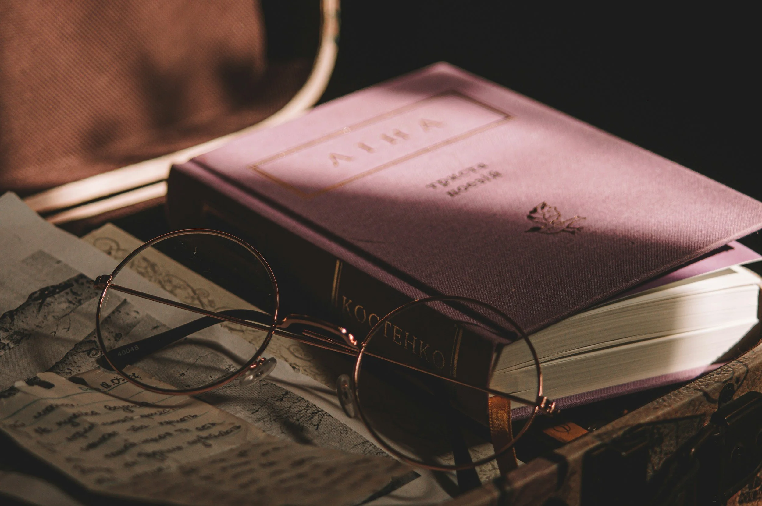 A close-up of a pink hardcover book resting next to wire-rimmed reading glasses on a vintage desk, demonstrating aesthetic website photos for literary authors
