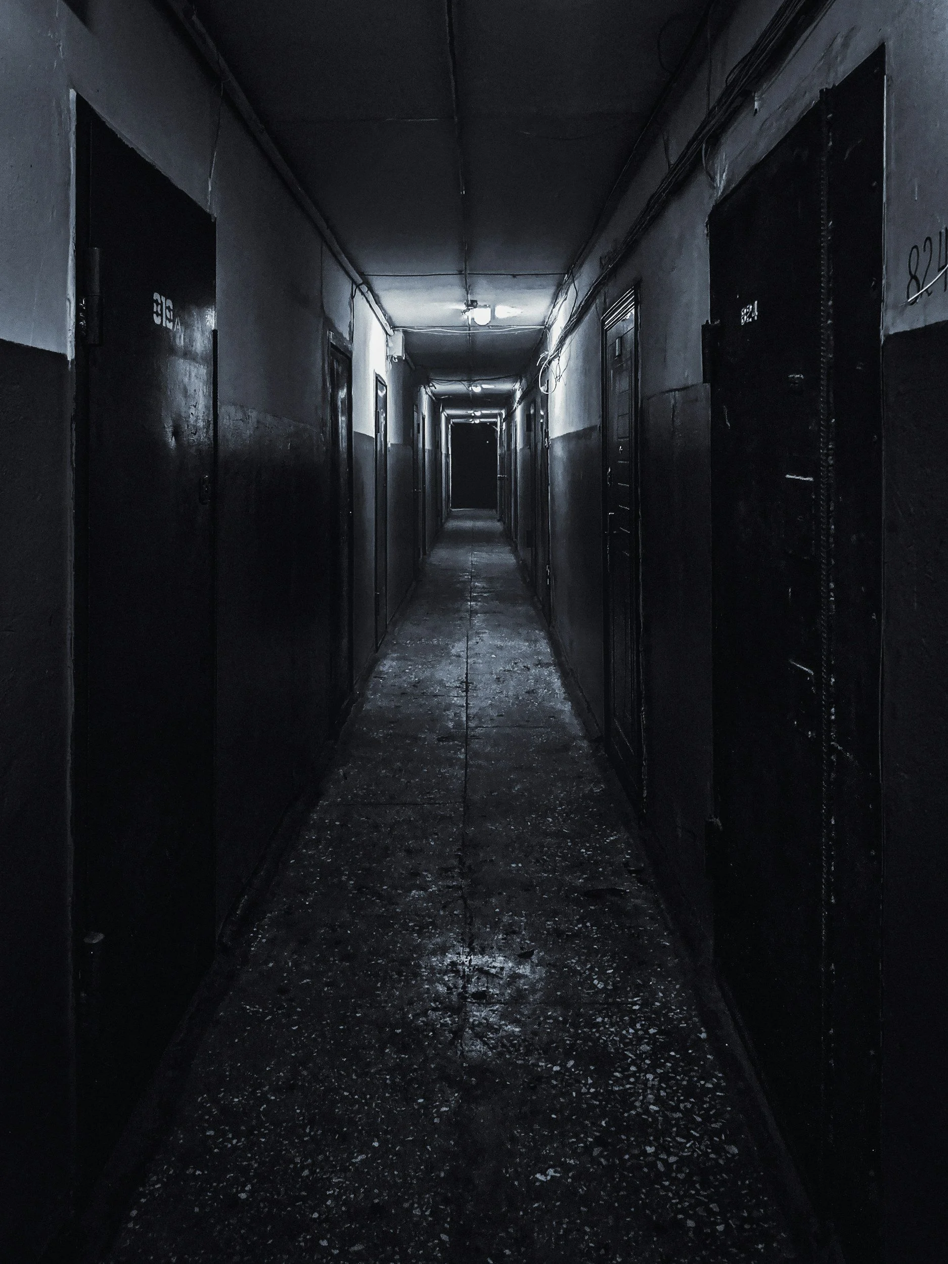 A stark, high-contrast perspective shot looking straight down a narrow, pitch-black institutional hallway with doors lining each side