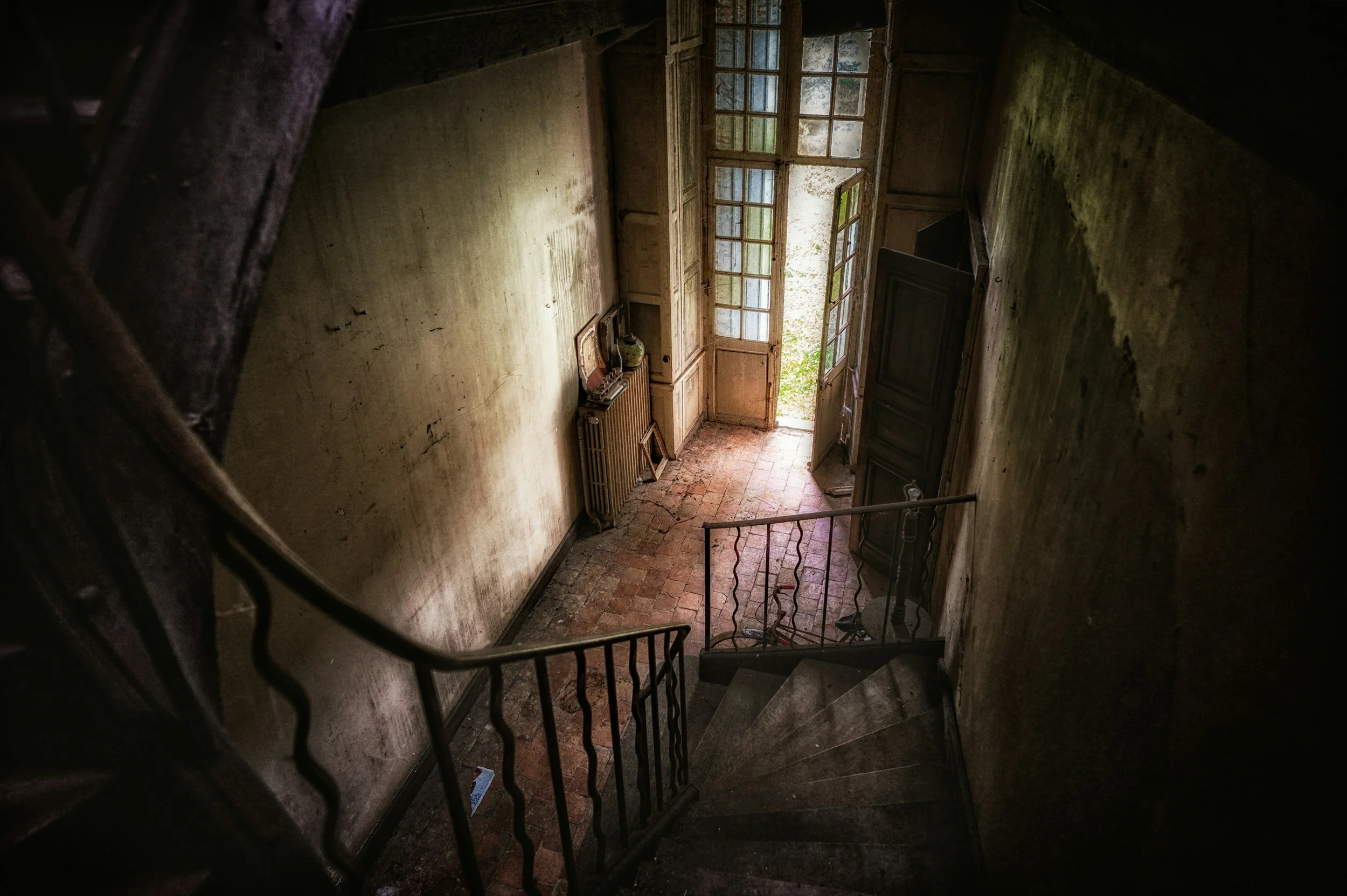 A dilapidated, peeling hallway in an old house with an ornate metal staircase railing